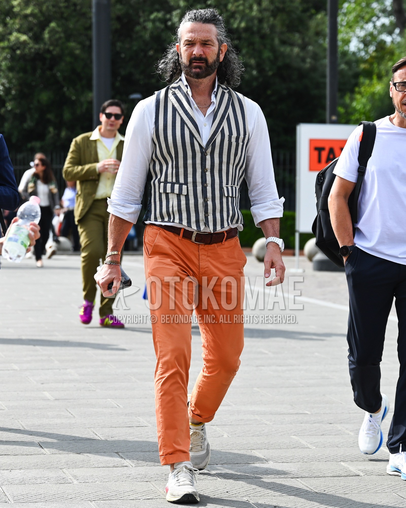 Men's spring summer autumn outfit with white black stripes gilet, white plain shirt, brown plain leather belt, orange plain chinos, white low-cut sneakers.