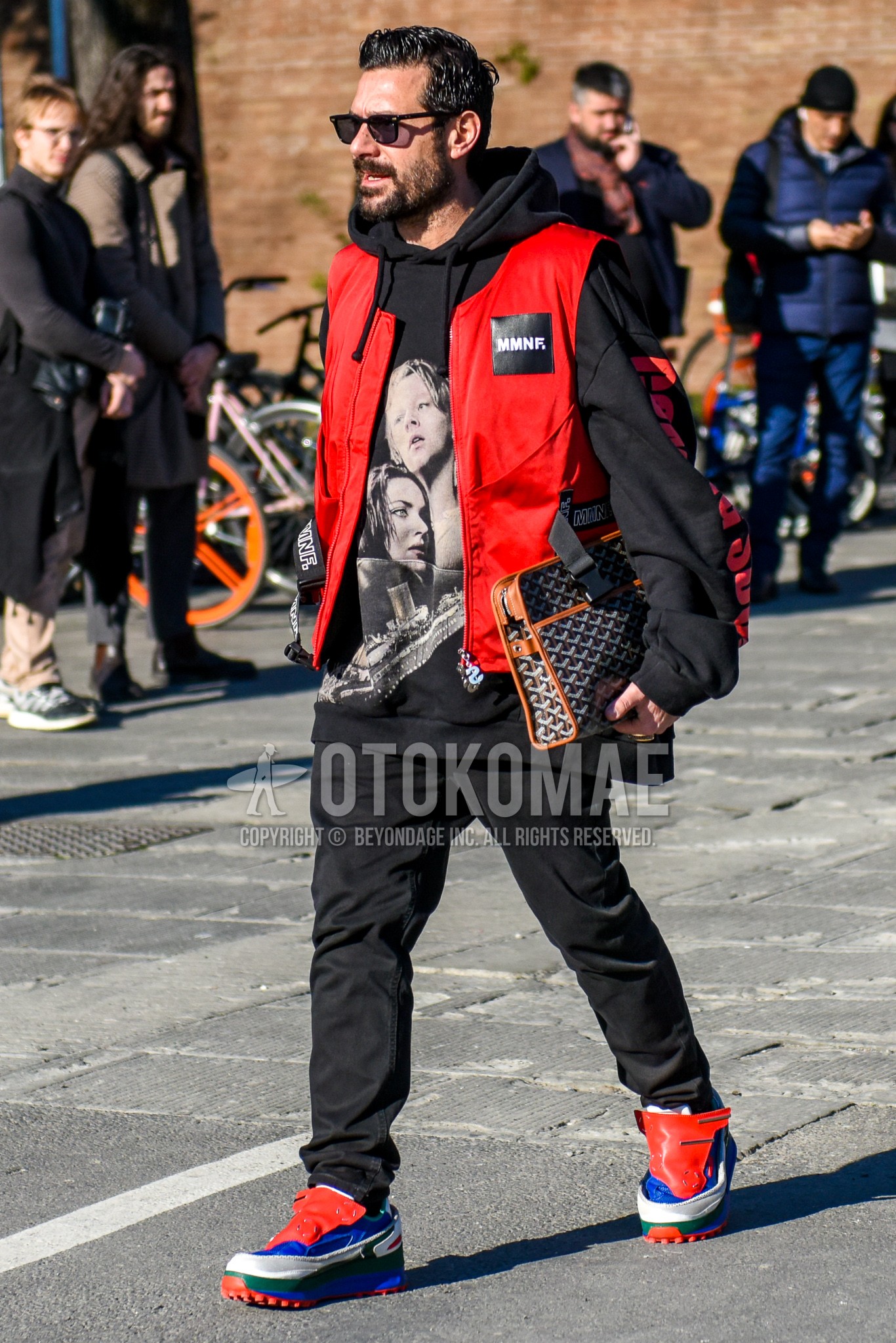 Men's autumn winter outfit with black plain sunglasses, black graphic hoodie, red one point casual vest, black plain denim/jeans, multi-color low-cut sneakers, multi-color bag clutch bag/second bag/drawstring bag.