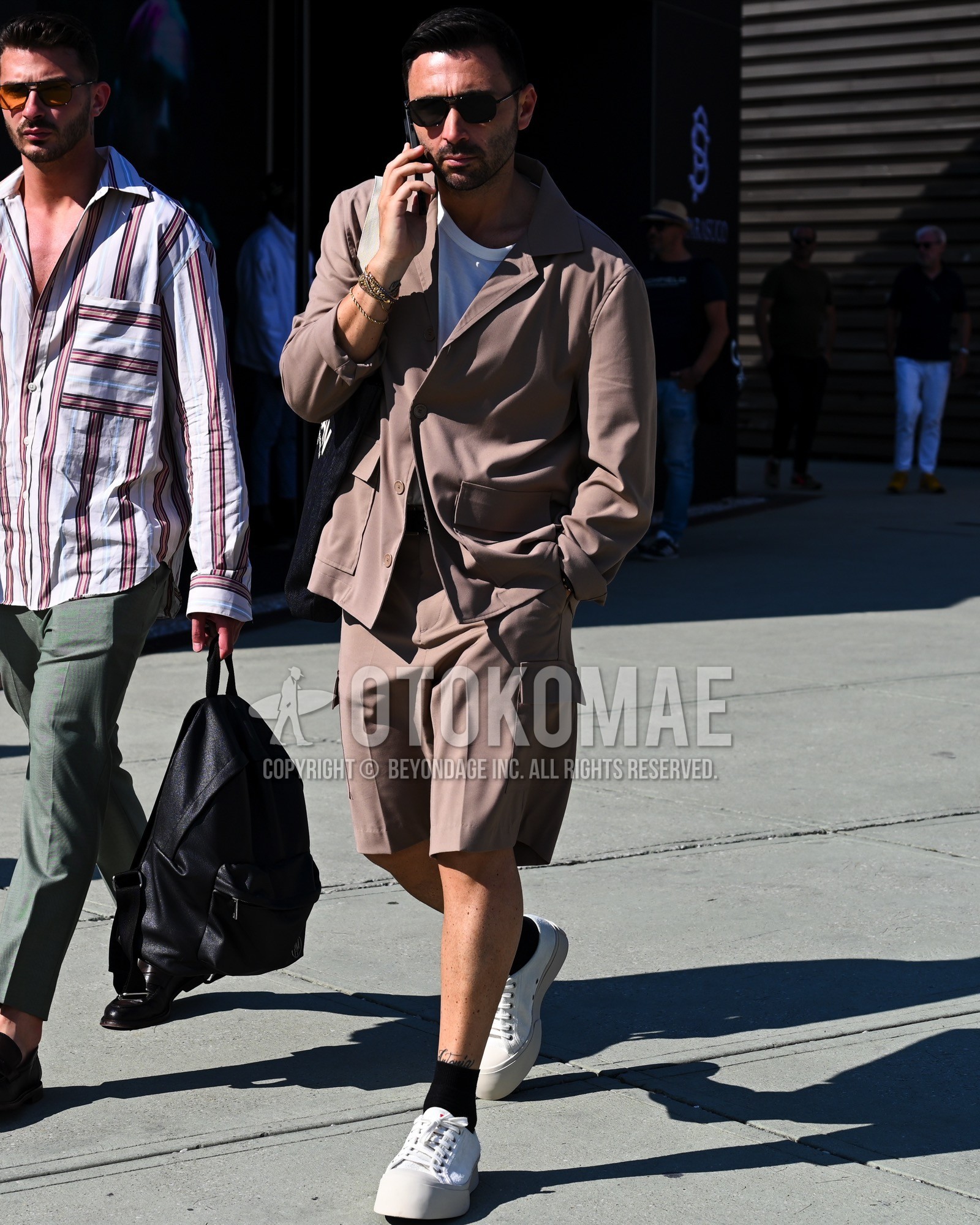 Men's spring summer outfit with black plain sunglasses, white plain t-shirt, black plain socks, white low-cut sneakers, beige plain casual setup.