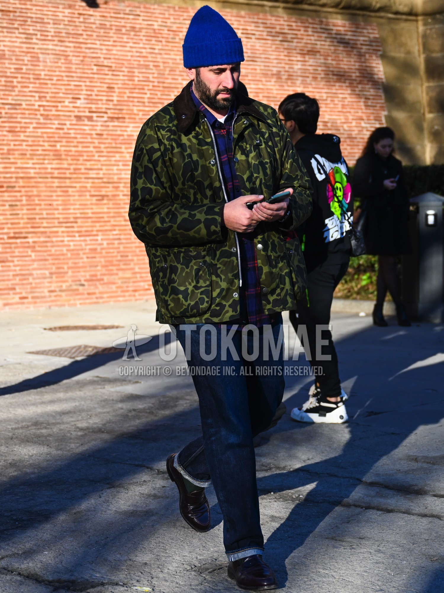 Men's autumn winter outfit with blue plain knit cap, olive green camouflage field jacket/hunting jacket, blue check shirt, white plain t-shirt, navy plain denim/jeans, green plain socks, brown coin loafers leather shoes.