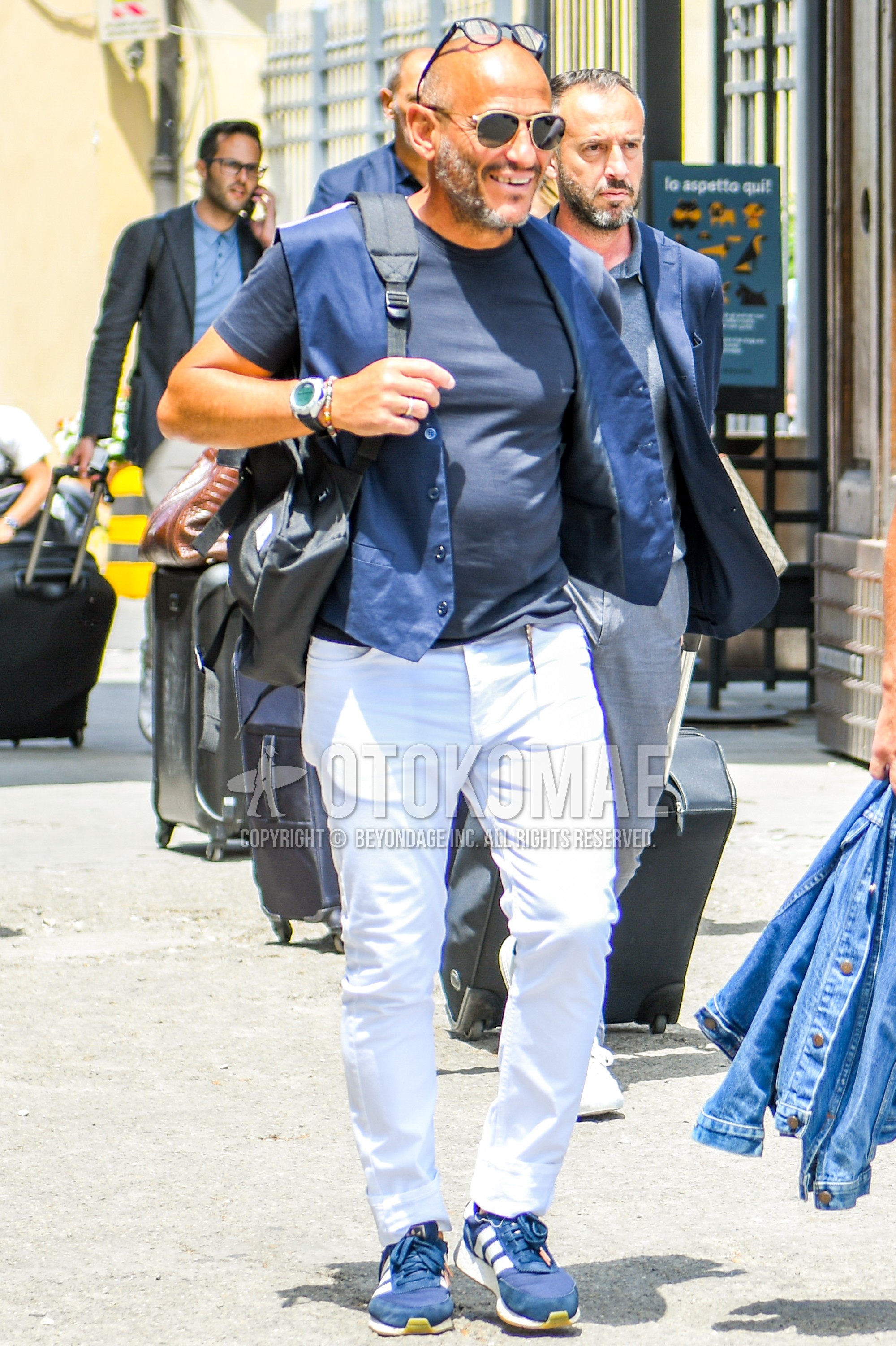 Men's summer outfit with plain sunglasses, blue plain gilet, navy plain t-shirt, white plain denim/jeans, navy low-cut sneakers.