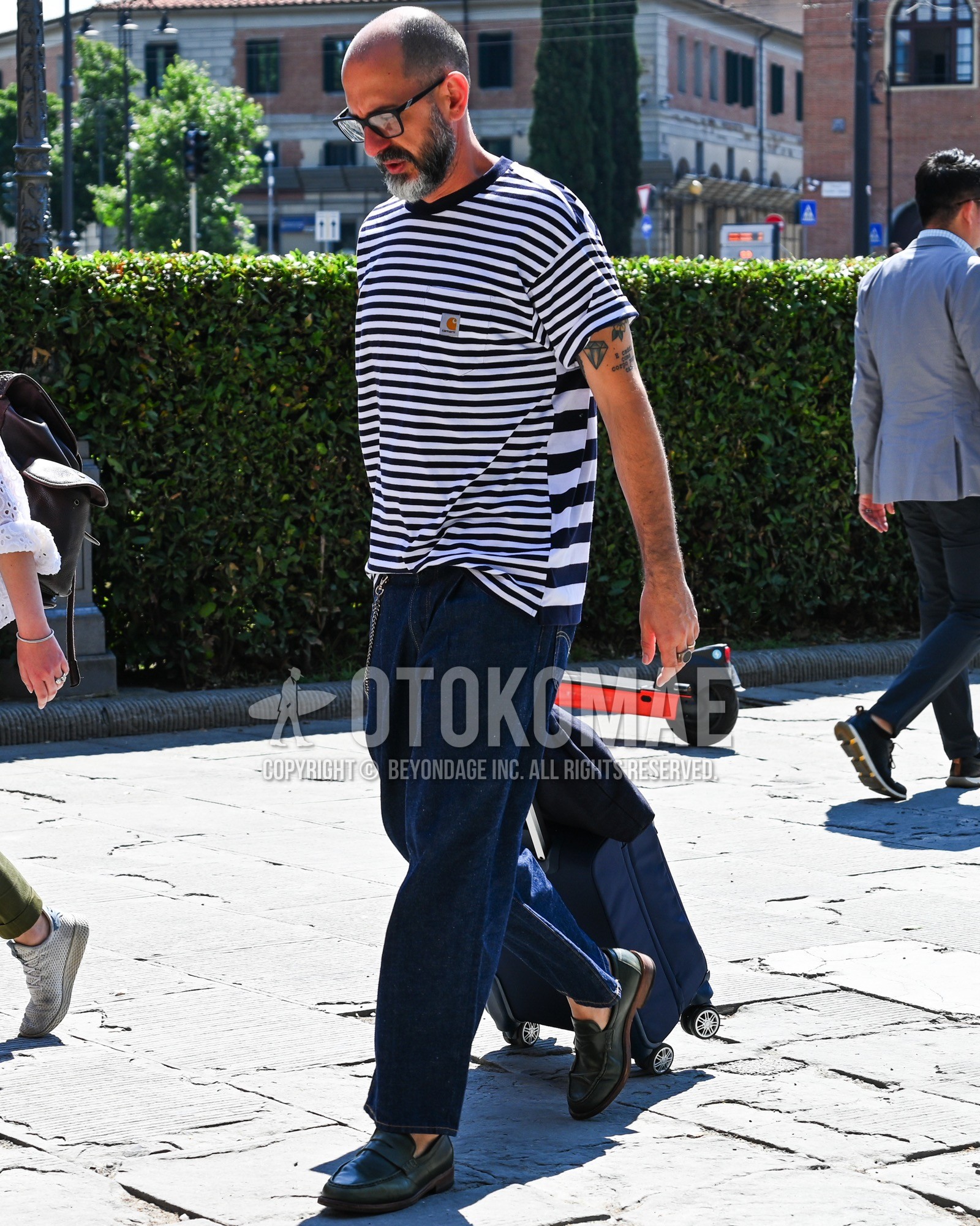 Men's spring summer outfit with clear plain sunglasses, navy white horizontal stripes t-shirt, blue plain denim/jeans, black coin loafers leather shoes, navy plain suitcase.