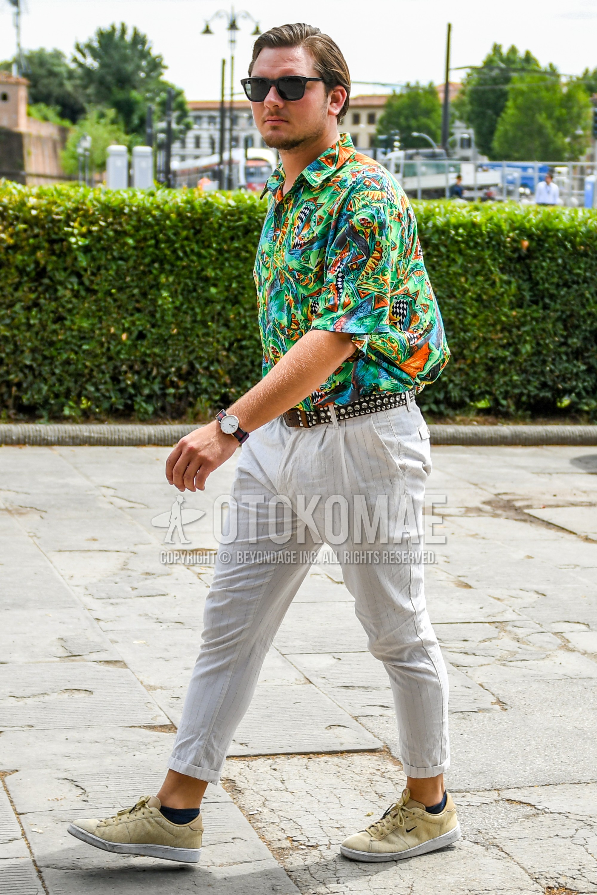Men's spring summer outfit with black plain sunglasses, green tops/innerwear shirt, brown belt leather belt, white stripes cotton pants, beige low-cut sneakers.