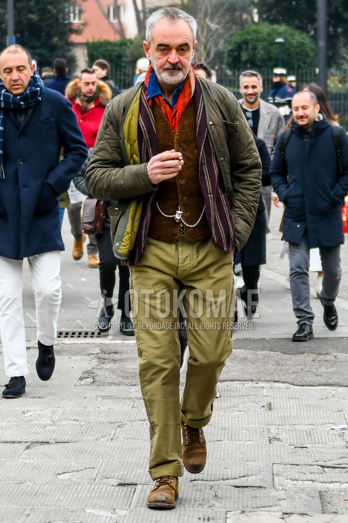 Men's winter outfit with red check scarf, olive green plain quilted jacket, brown plain gilet, olive green plain cotton pants, brown work boots.