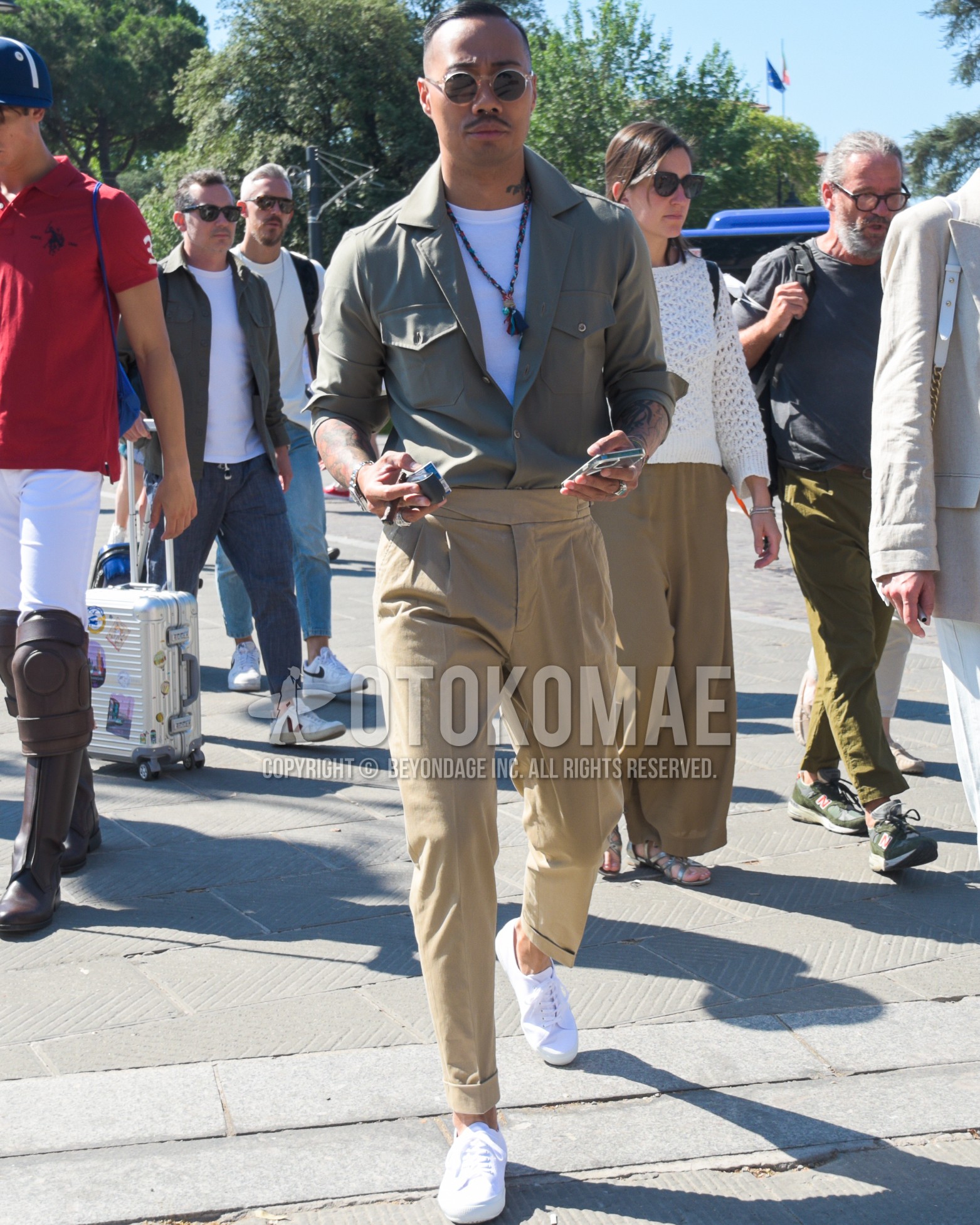 Men's spring summer outfit with brown clear plain sunglasses, olive green plain shirt, white plain t-shirt, beige plain beltless pants, white low-cut sneakers.
