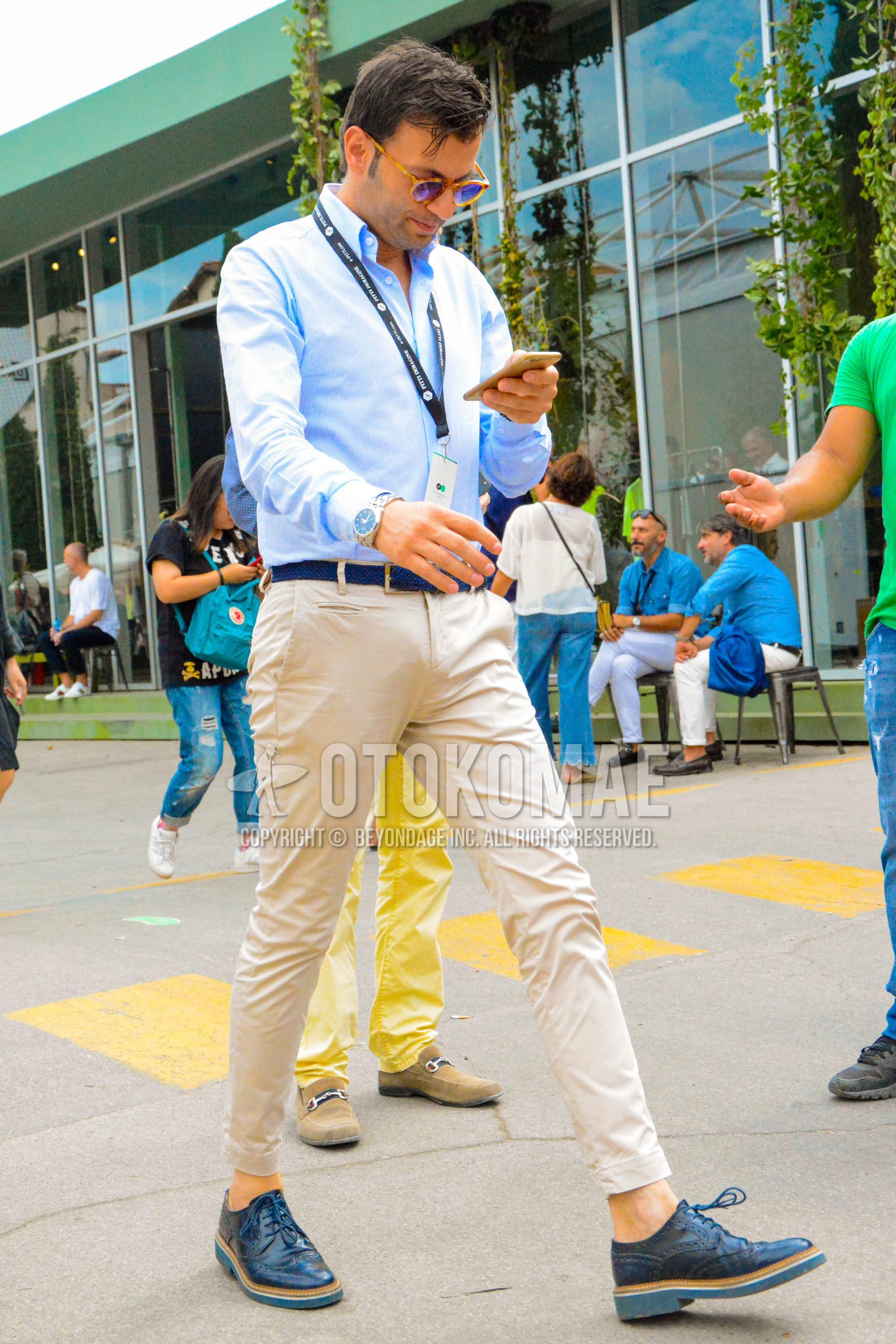 Men's spring summer outfit with beige tortoiseshell sunglasses, light blue plain shirt, blue plain braided belt, beige plain chinos, beige plain ankle pants, navy brogue shoes leather shoes.