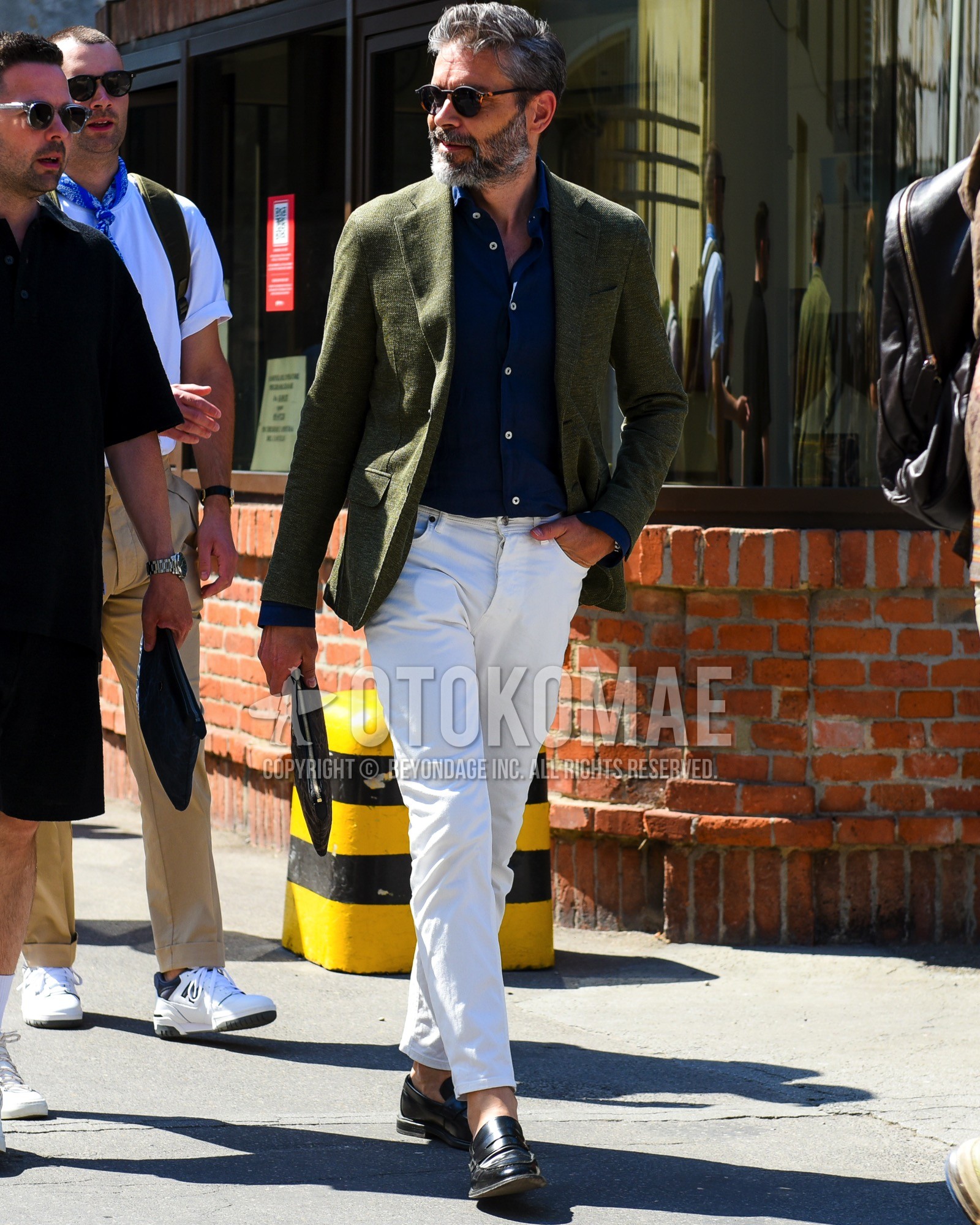 Men's spring summer outfit with brown tortoiseshell sunglasses, olive green plain tailored jacket, navy plain shirt, white plain cotton pants, black coin loafers leather shoes, black bag clutch bag/second bag/drawstring bag.