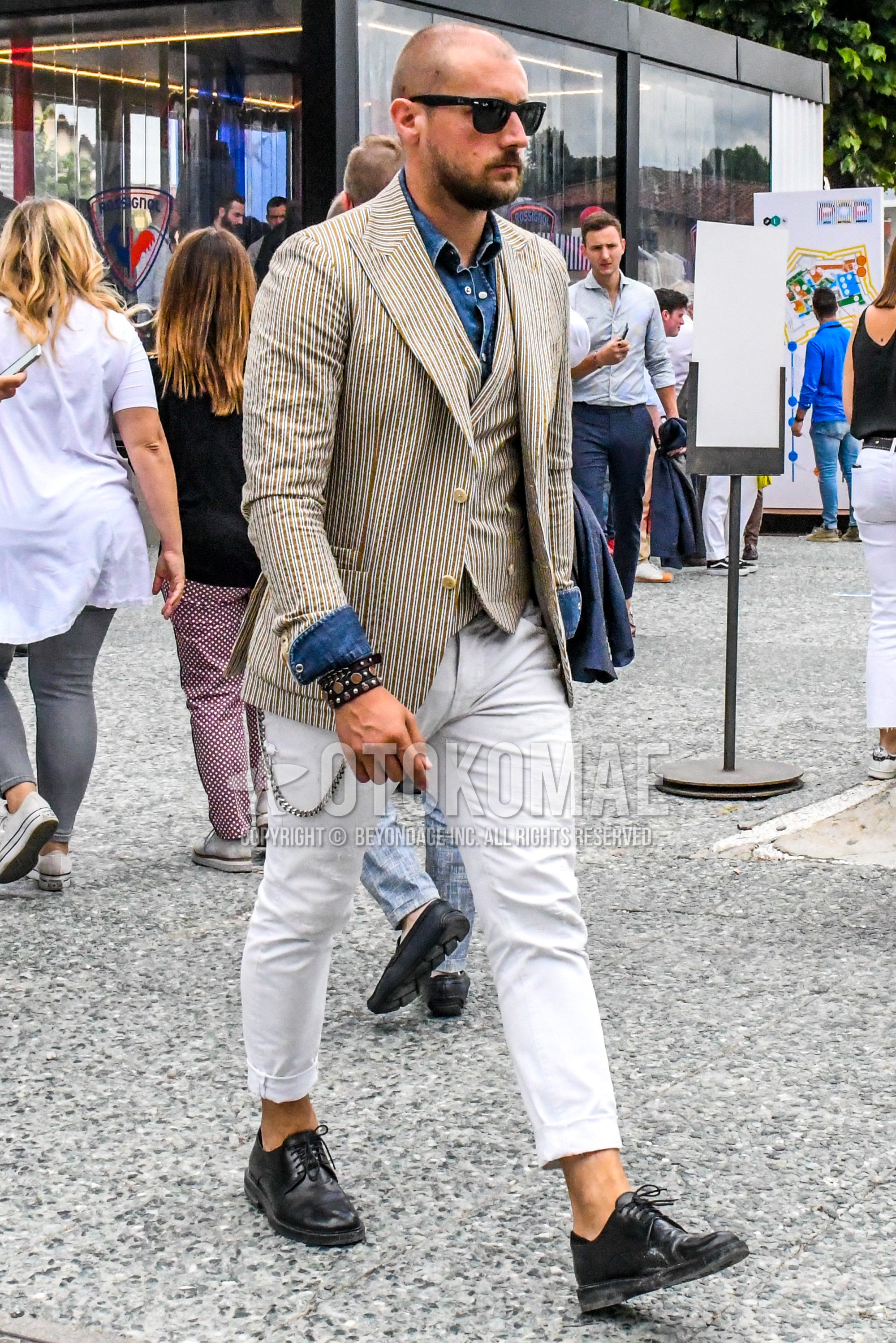 Men's winter outfit with plain sunglasses, beige stripes tailored jacket, beige stripes gilet, blue plain denim shirt/chambray shirt, white plain cotton pants, black plain toe leather shoes.