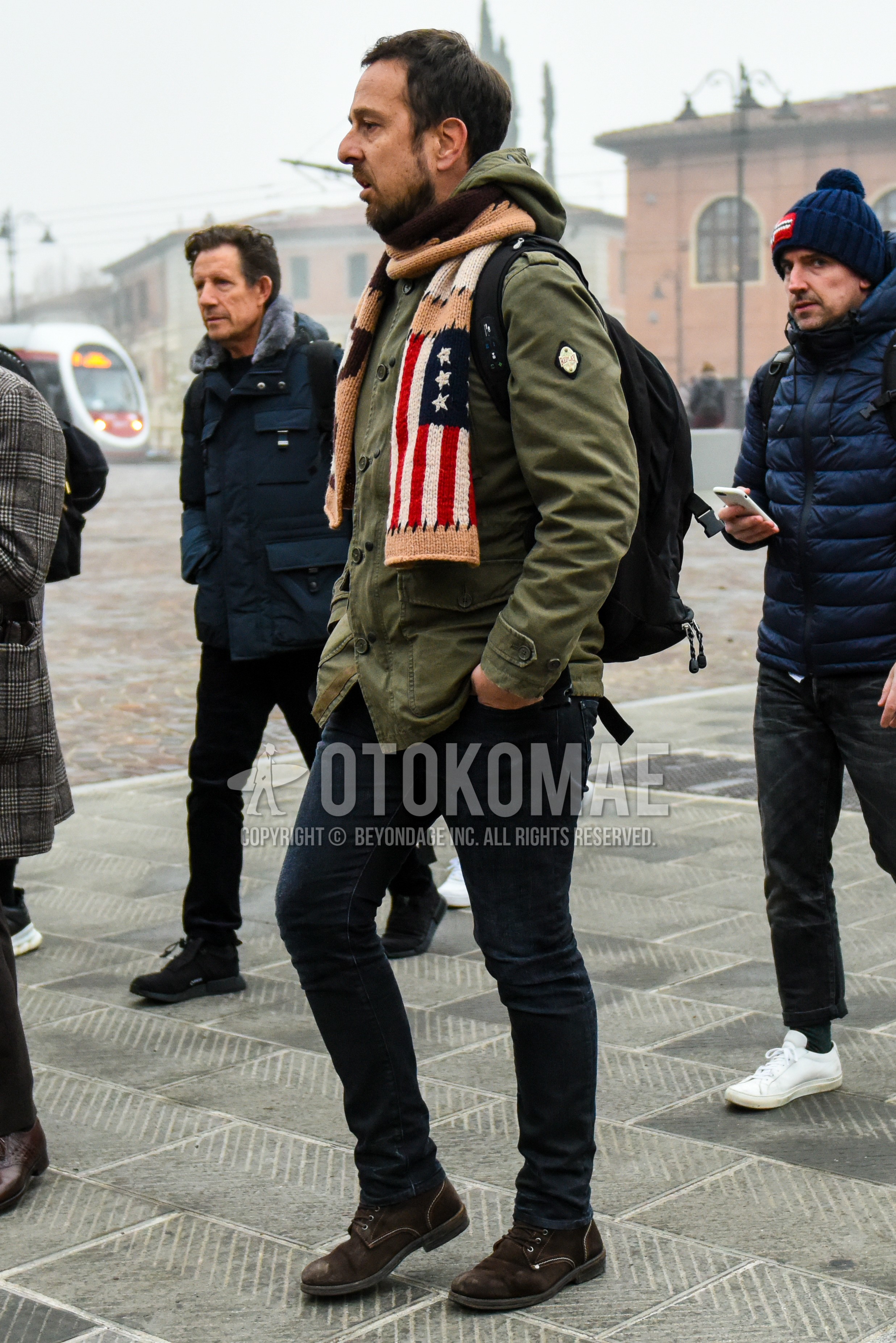 Men's winter outfit with multi-color graphic scarf, olive green plain hooded coat, black plain denim/jeans, brown  boots.
