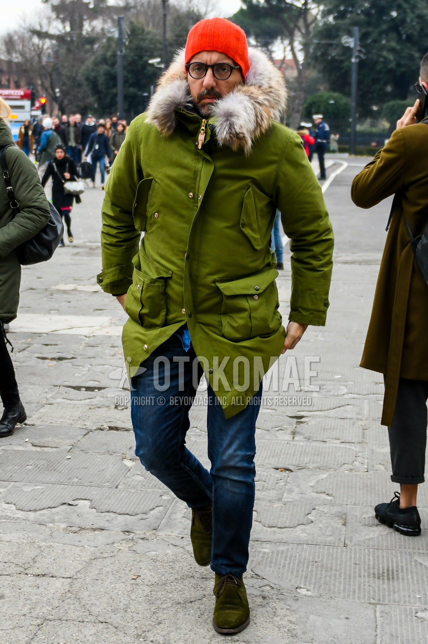 Men's winter outfit with orange plain knit cap, plain glasses, green plain mod coat, blue plain denim/jeans, olive green chukka boots.