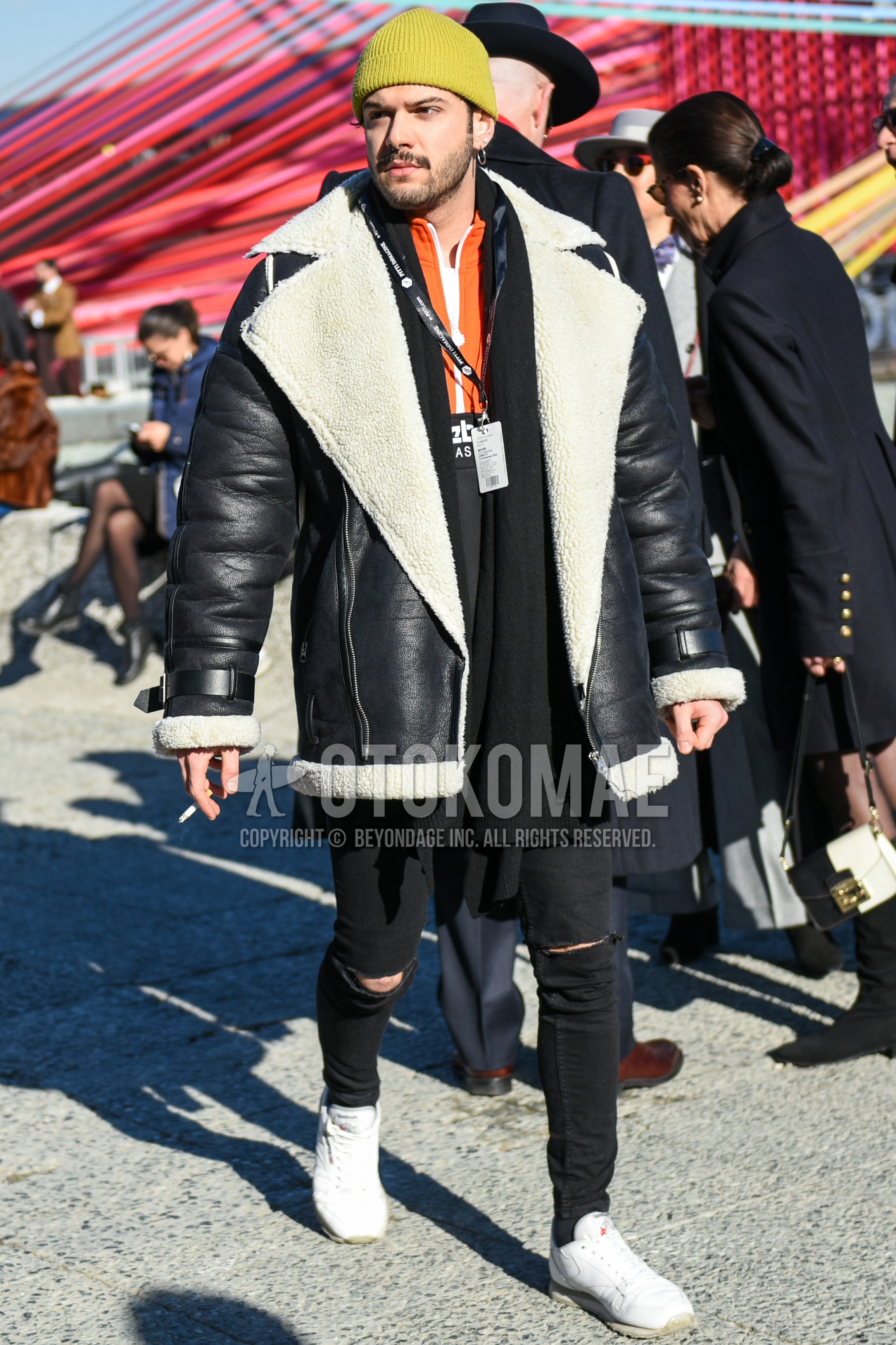 Men's autumn winter outfit with yellow plain knit cap, black plain scarf, black plain leather jacket, orange black plain windbreaker, black plain damaged jeans, white low-cut sneakers.