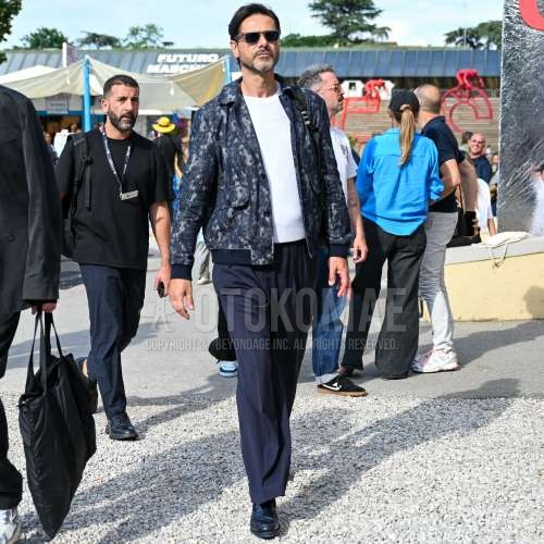 クリア無地のサングラス、ネイビーアウター、白無地のTシャツ、ネイビー無地のスラックス、ネイビー無地のプリーツパンツ、黒コインローファーの革靴、黒無地のバックパックを合わせた春夏のメンズコーデ・着こなし。