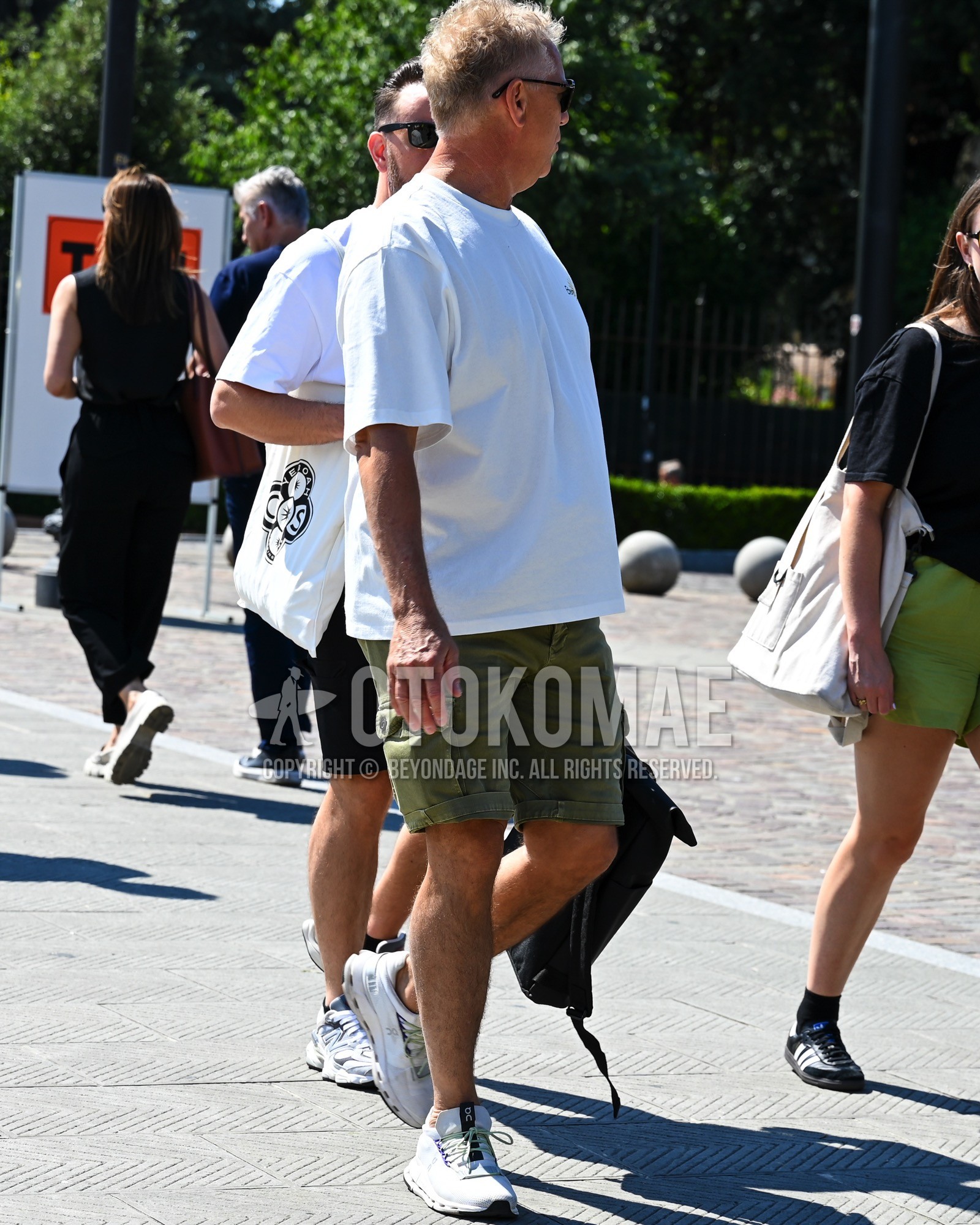 Men's spring summer outfit with black plain sunglasses, white plain t-shirt, olive green plain short pants, white low-cut sneakers, black plain backpack.