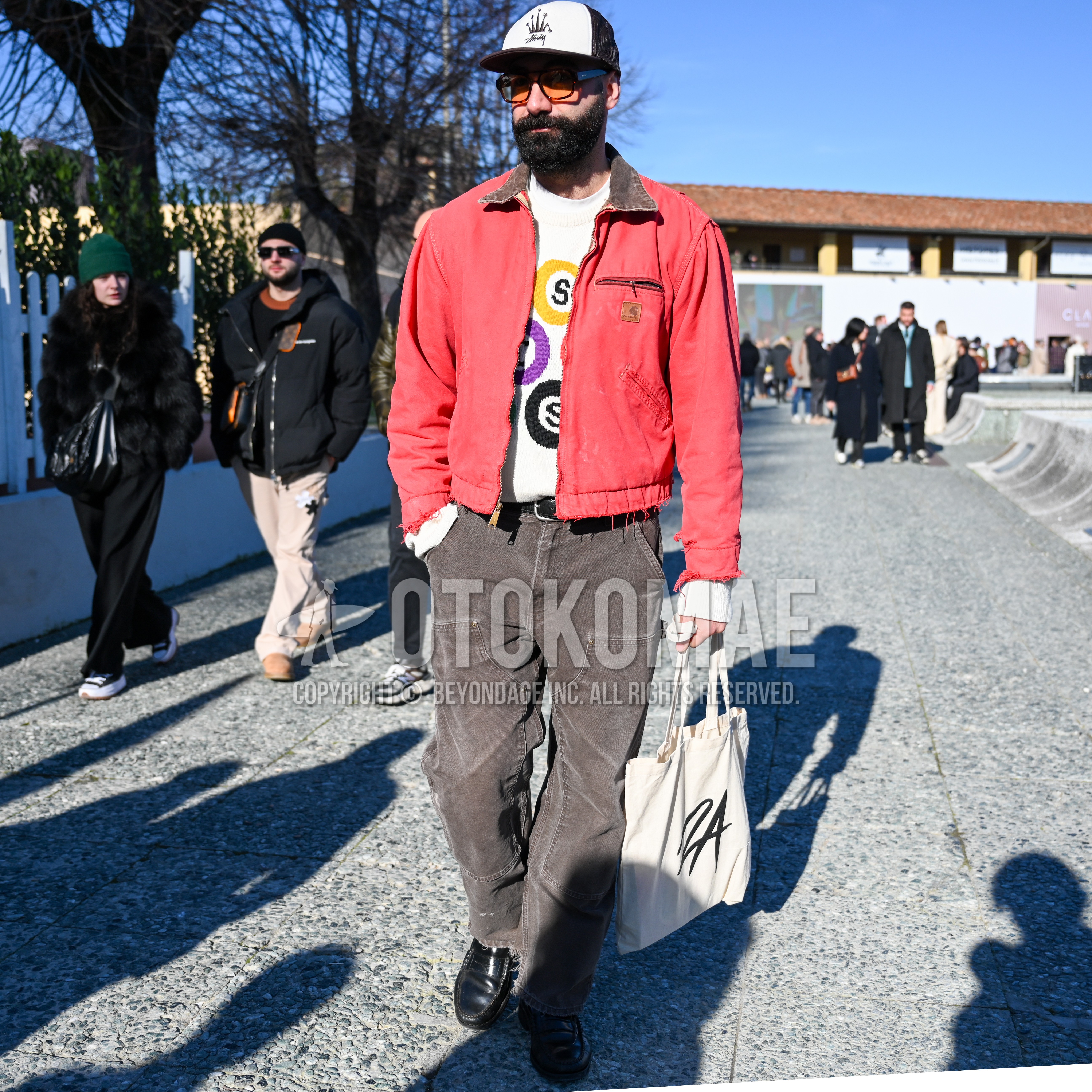 Men's spring autumn outfit with black one point baseball cap, brown tortoiseshell sunglasses, red plain outerwear, white graphic sweater, brown plain work pants, black coin loafers leather shoes, beige one point tote bag.