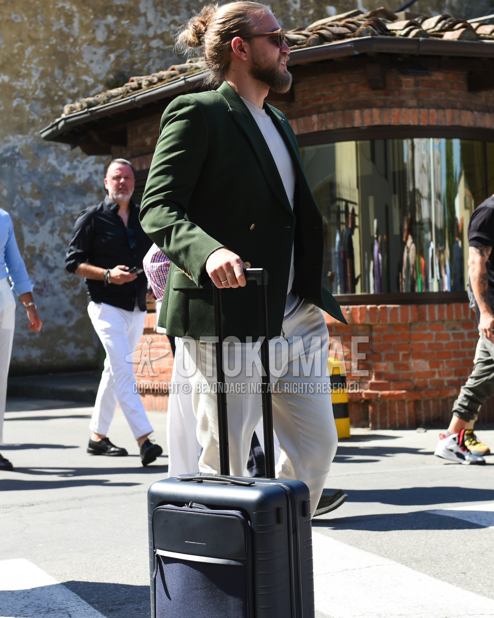 Men's spring summer outfit with brown plain sunglasses, olive green plain tailored jacket, white plain t-shirt, white bottoms cotton pants, black plain suitcase.