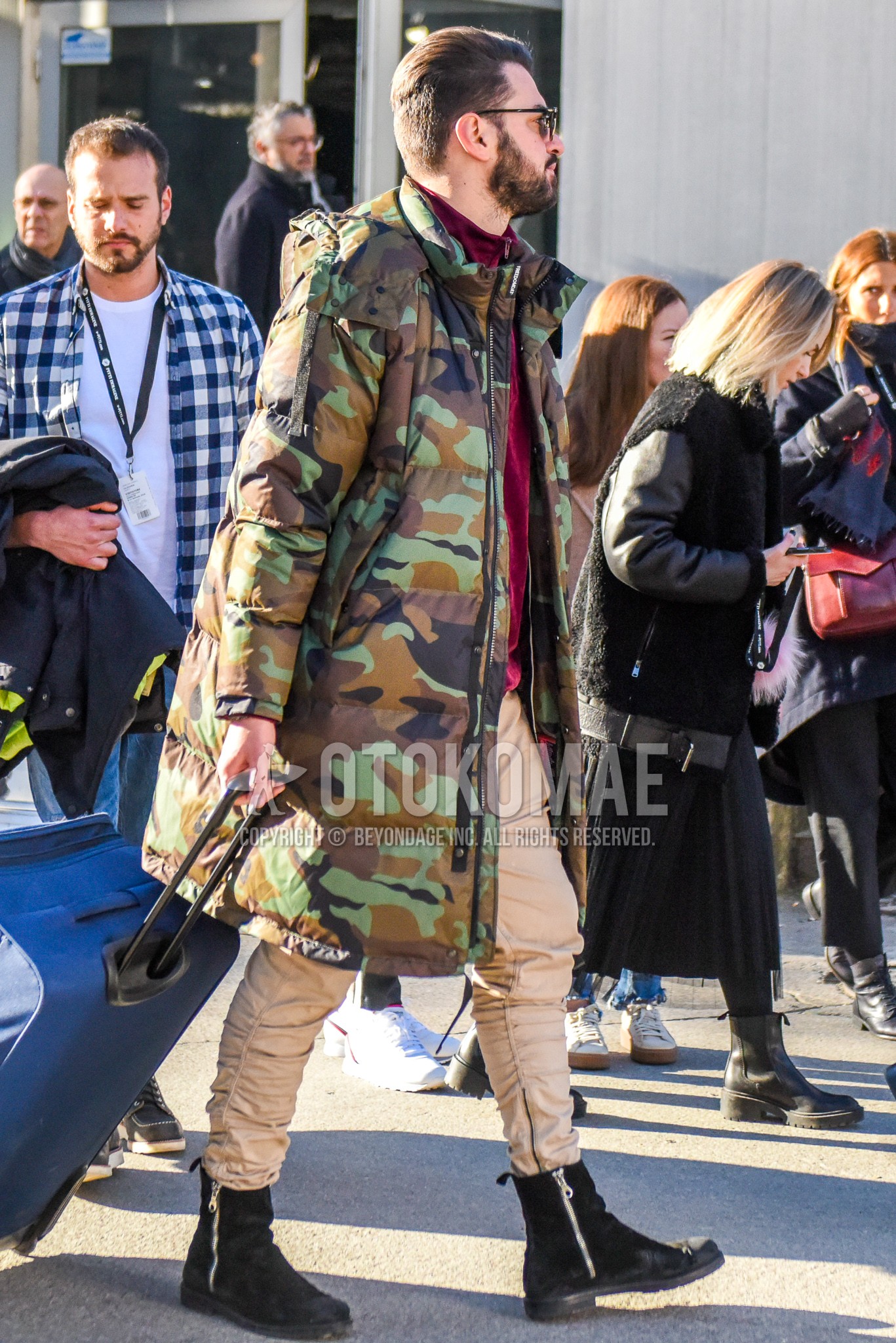 Men's winter outfit with black plain sunglasses, olive green brown camouflage down jacket, red plain cardigan, beige plain chinos, black  boots, blue plain suitcase.