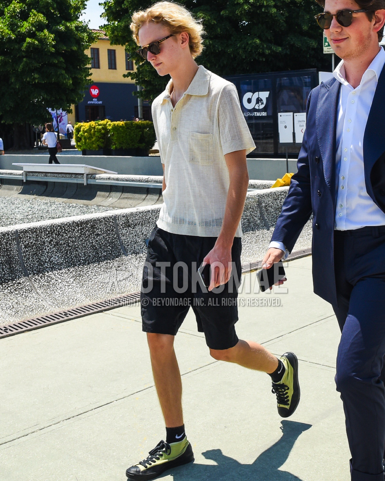 Men's spring summer outfit with brown tortoiseshell sunglasses, beige plain polo shirt, black plain short pants, black one point socks, yellow low-cut sneakers.