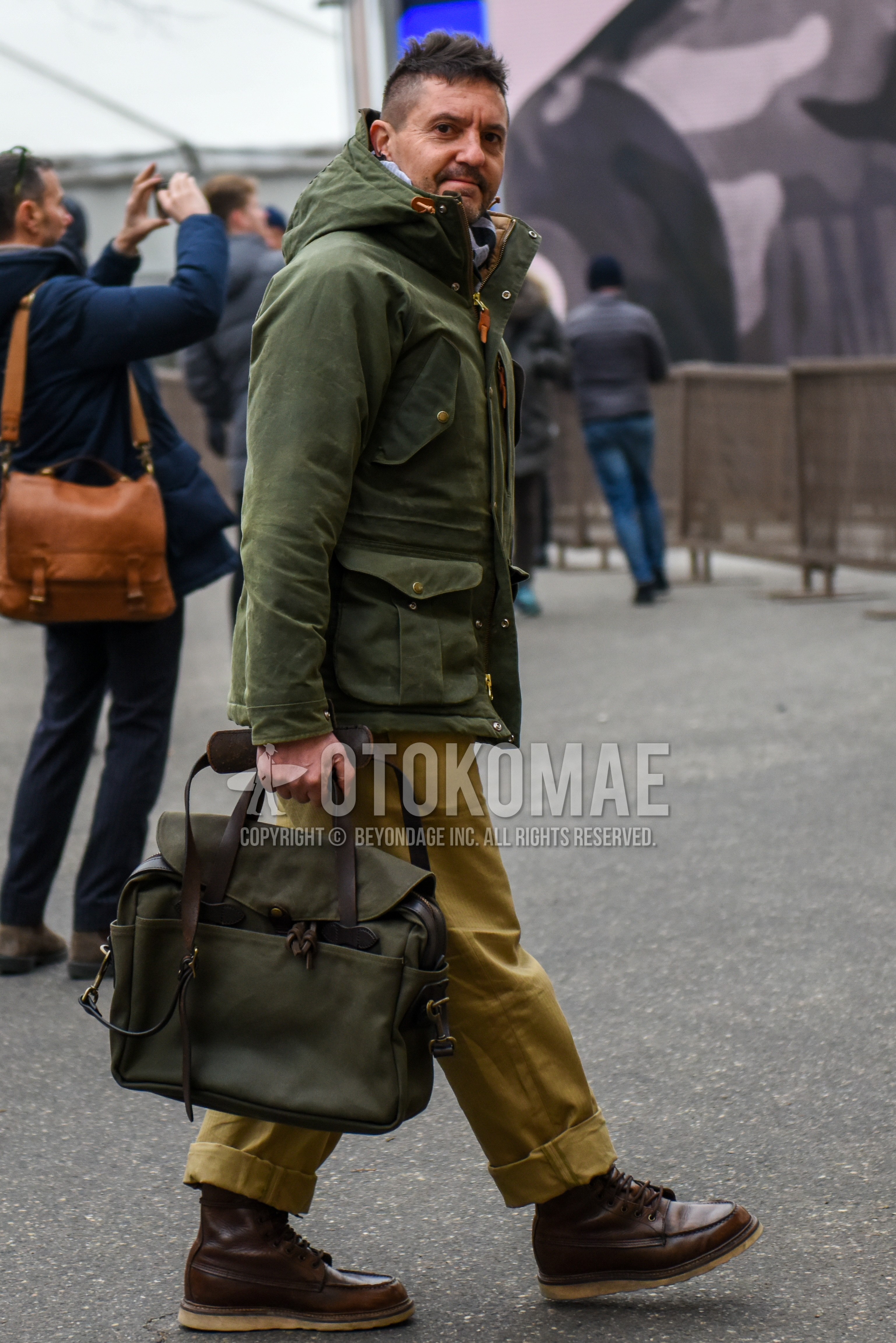 Men's winter outfit with olive green plain hooded coat, beige plain chinos, beige plain wide pants, brown work boots, olive green plain briefcase/handbag.