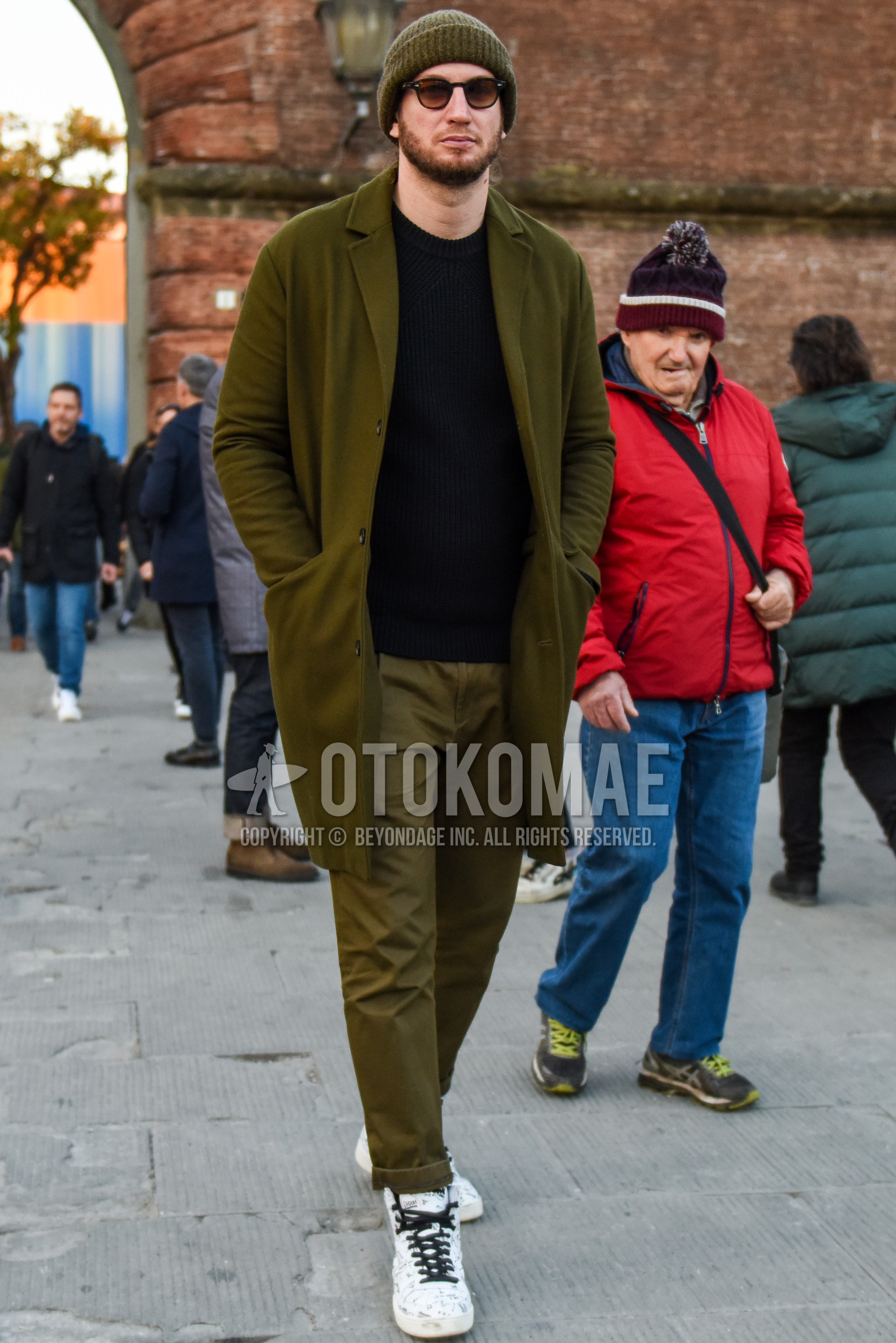 Men's autumn winter outfit with olive green plain knit cap, black plain sunglasses, olive green plain chester coat, black plain sweater, olive green plain chinos, white high-cut sneakers.