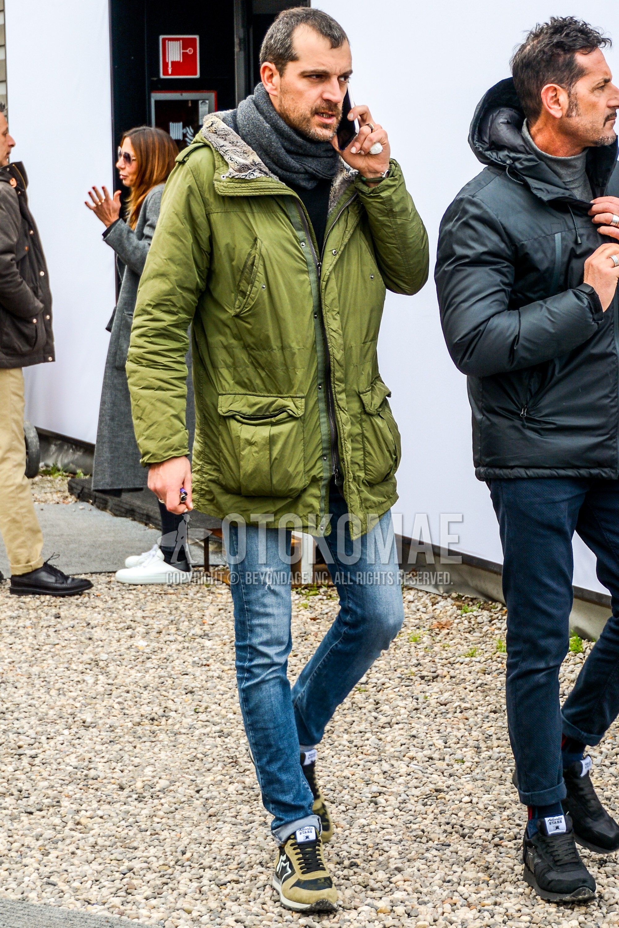Men's winter outfit with gray plain scarf, olive green plain hooded coat, blue plain denim/jeans, beige low-cut sneakers.