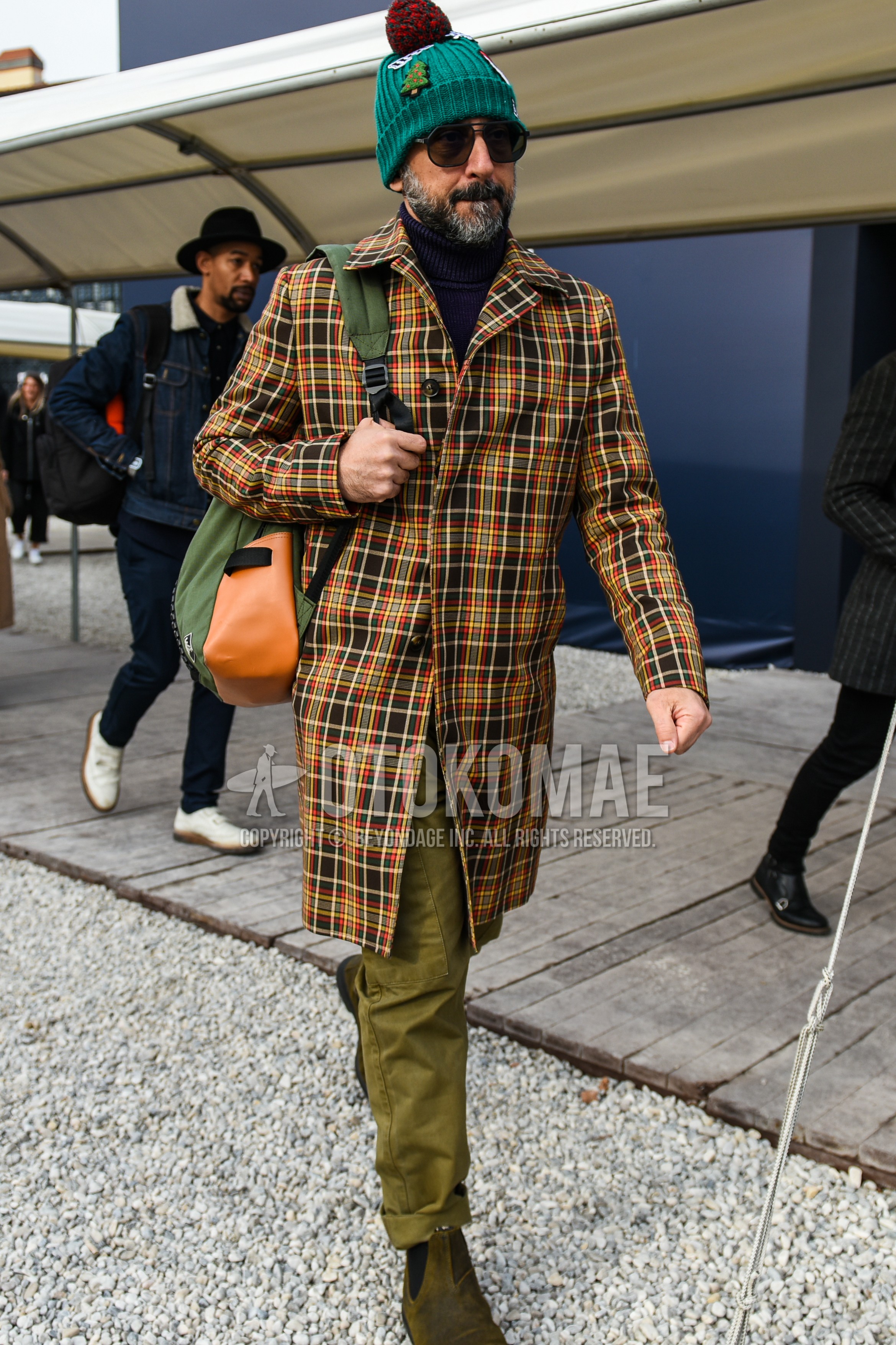 Men's autumn winter outfit with green plain knit cap, brown plain sunglasses, multi-color check stenkarrer coat, black plain turtleneck knit, olive green plain chinos, brown side-gore boots, olive green plain backpack.