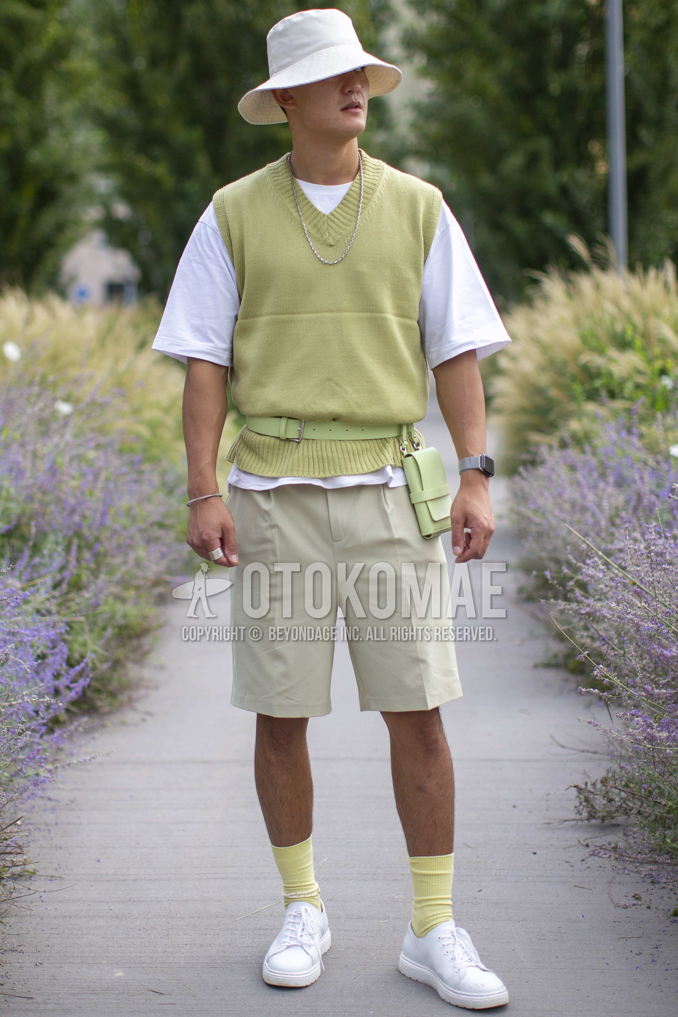 Men's spring summer outfit with white plain bucket hat, green plain casual vest, white plain t-shirt, beige plain short pants, yellow plain socks, white sneakers, green plain body bag.