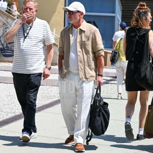 白無地のベースボールキャップ、黒無地のサングラス、ベージュ無地のシャツジャケット、ニットの白無地のポロシャツ、白無地のスラックス、ブラウンローカットのスニーカー、黒無地のバックパックを合わせた春夏のメンズコーデ・着こなし。