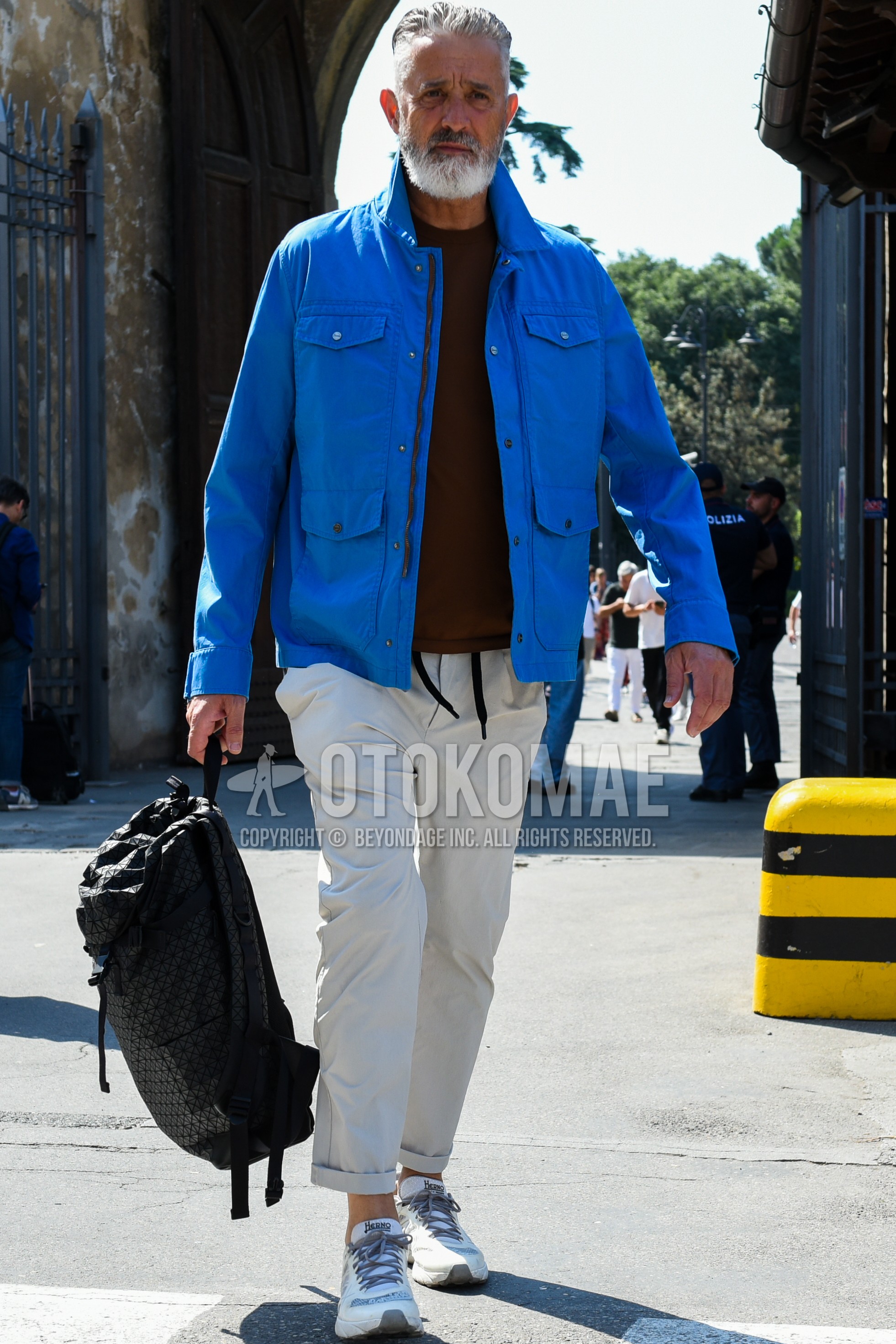 Men's spring summer outfit with blue plain shirt jacket, red plain t-shirt, beige plain ankle pants, beige plain easy pants, white low-cut sneakers, black plain backpack.