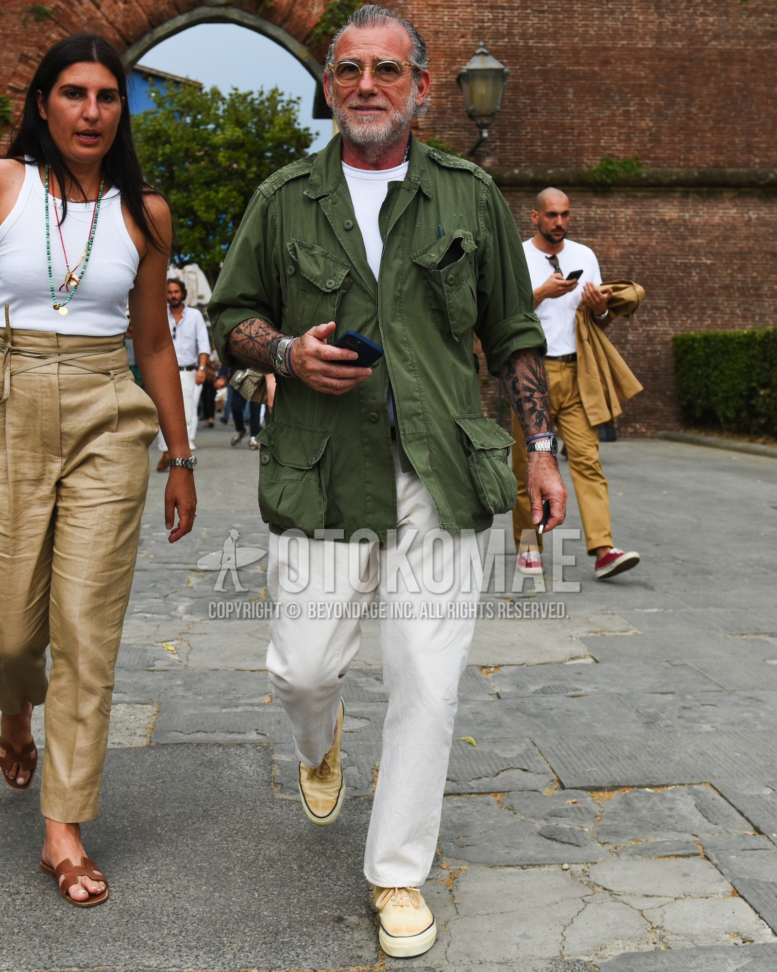 Men's spring summer outfit with silver plain sunglasses, olive green plain field jacket/hunting jacket, white plain t-shirt, white plain cotton pants, beige low-cut sneakers.