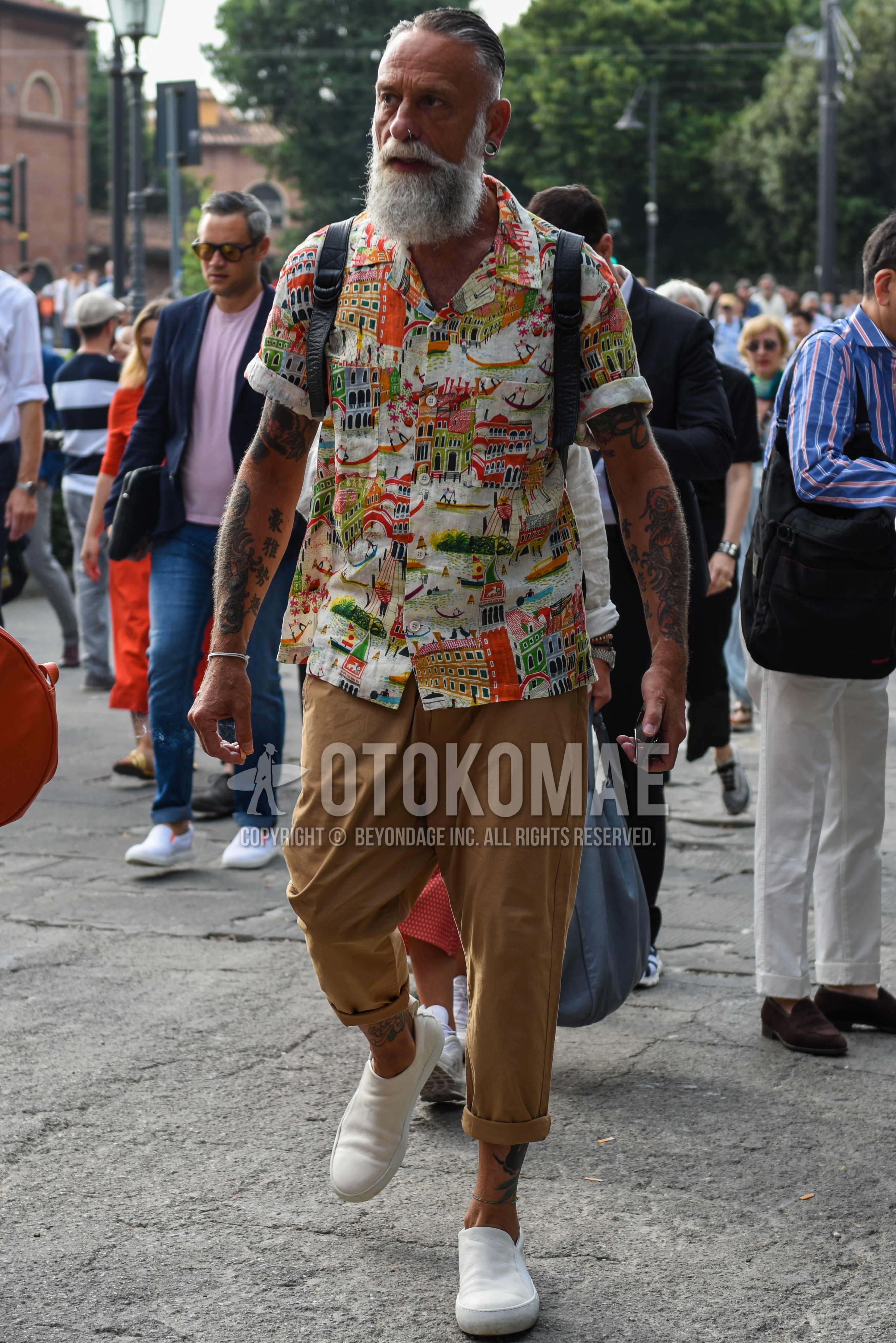 Men's summer outfit with multi-color graphic shirt, beige plain cotton pants, white slip-on sneakers.