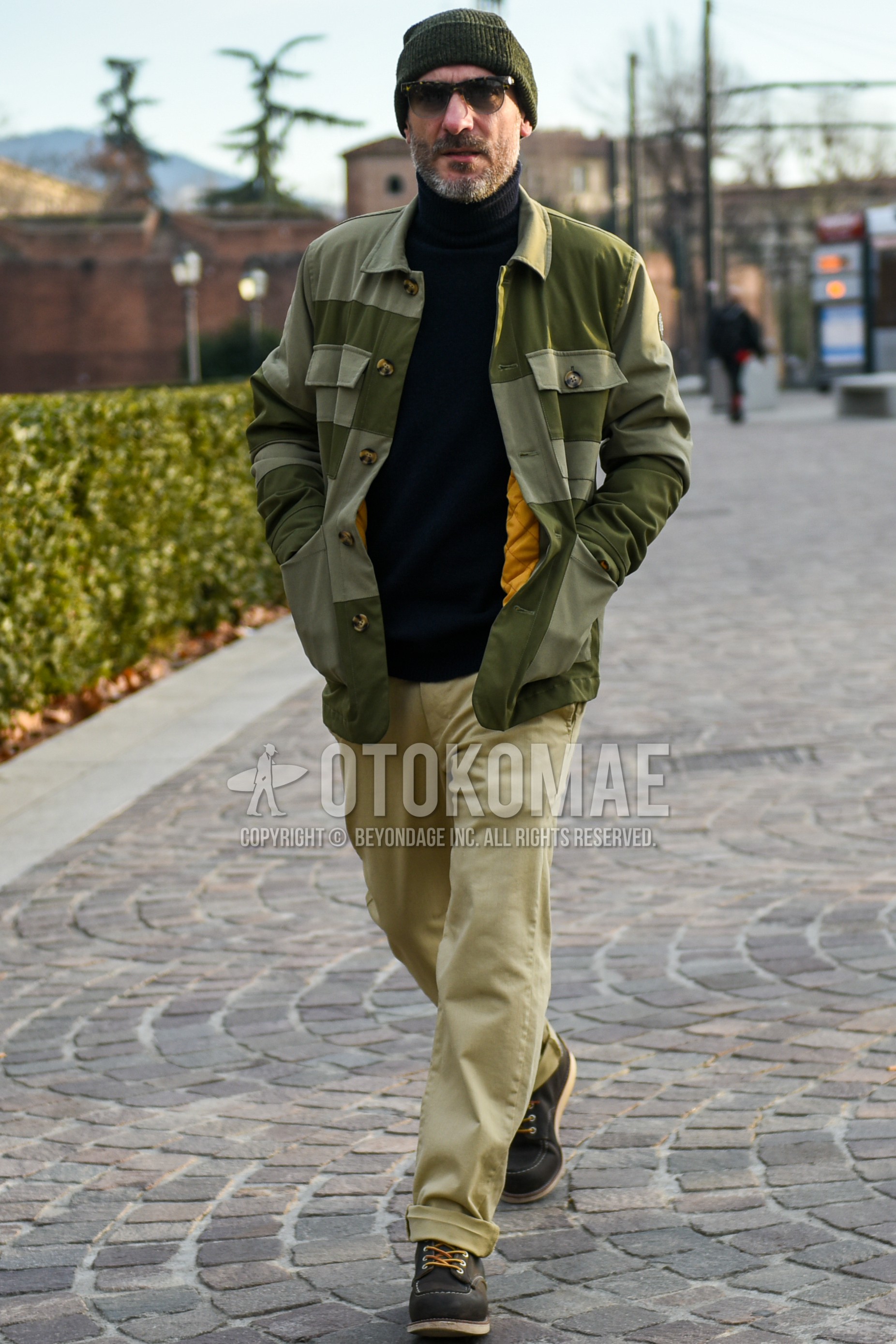 Men's autumn winter outfit with olive green plain knit cap, black plain sunglasses, olive green plain shirt jacket, black plain turtleneck knit, beige plain chinos, black work boots.