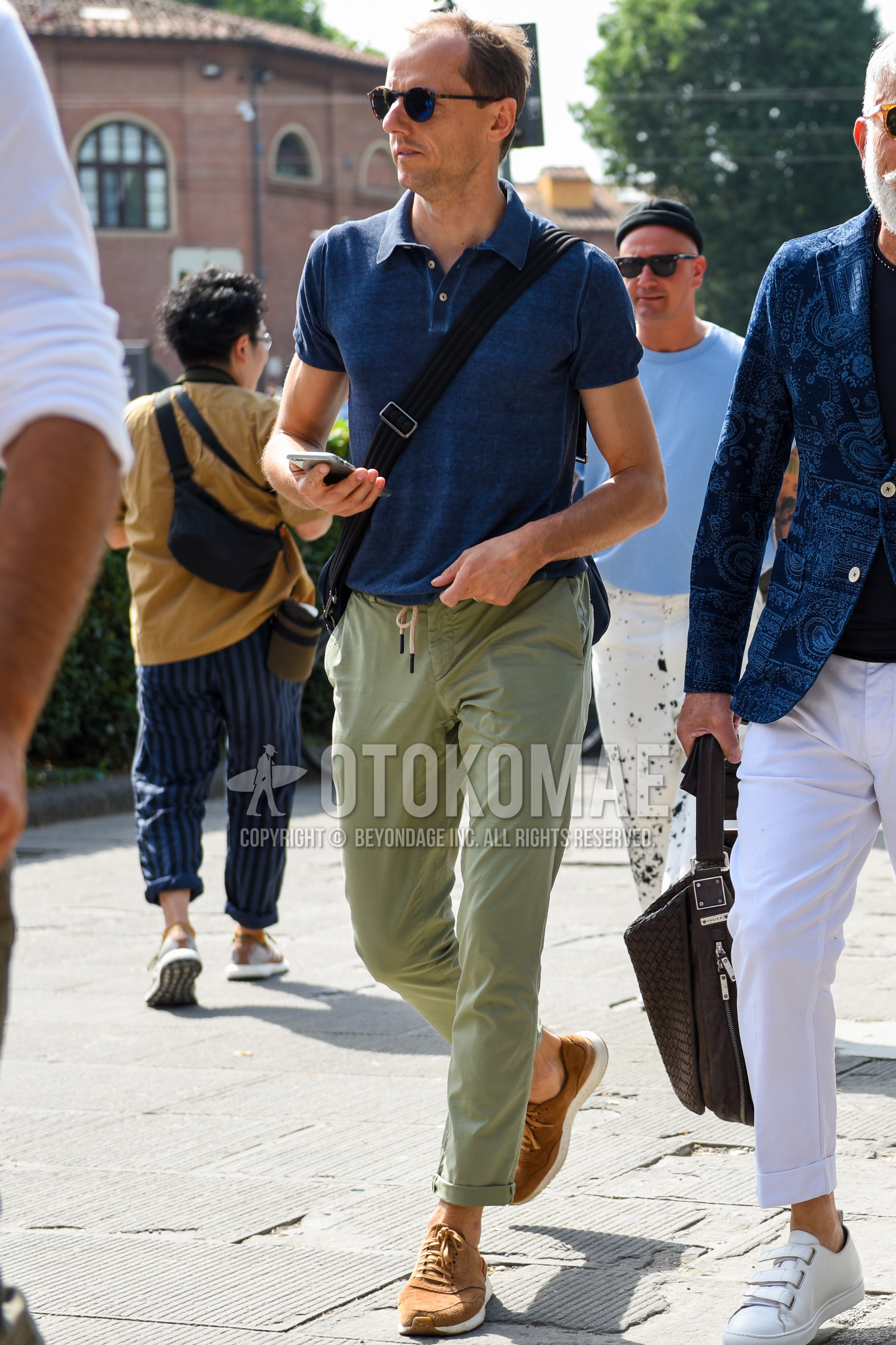 Men's spring summer outfit with beige tortoiseshell sunglasses, blue plain polo shirt, olive green plain easy pants, brown low-cut sneakers, black plain shoulder bag.