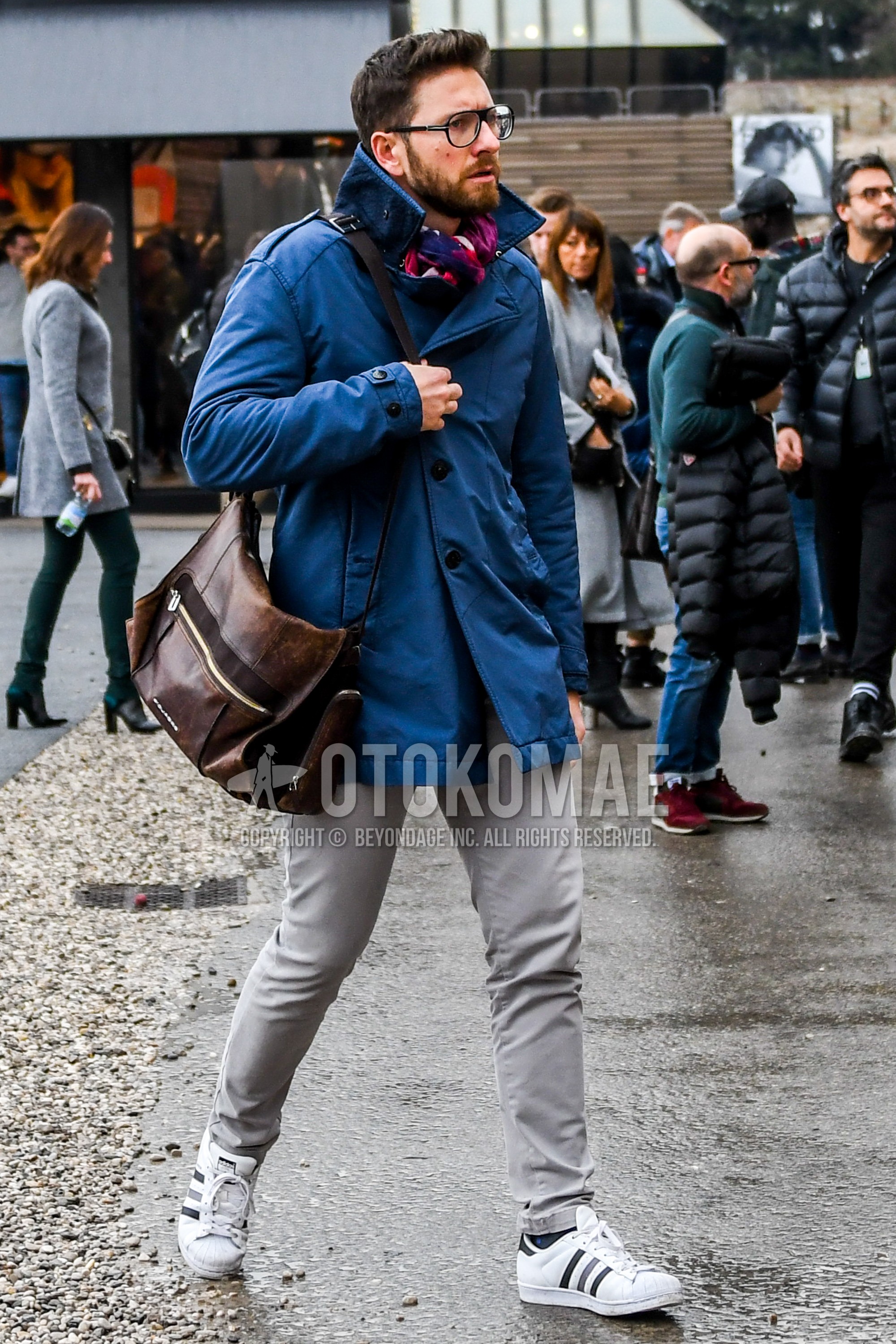 Men's autumn winter outfit with black plain glasses, multi-color scarf scarf, blue plain trench coat, gray plain chinos, white low-cut sneakers, brown plain shoulder bag.