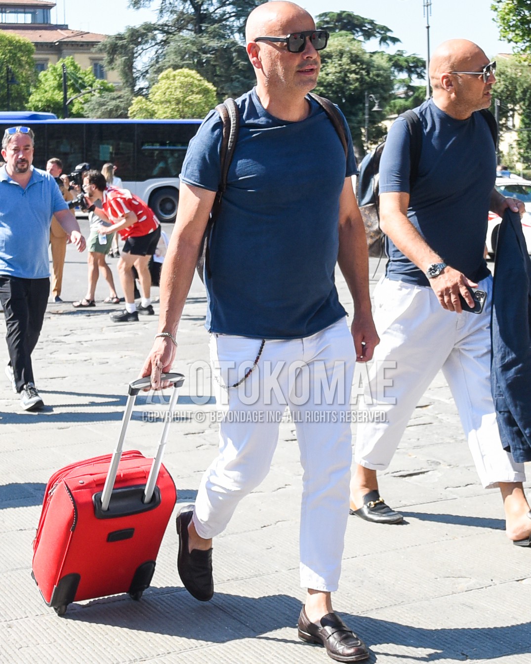 Men's spring summer outfit with black plain sunglasses, navy plain t-shirt, white plain cotton pants, brown coin loafers leather shoes, red plain suitcase, brown plain backpack.