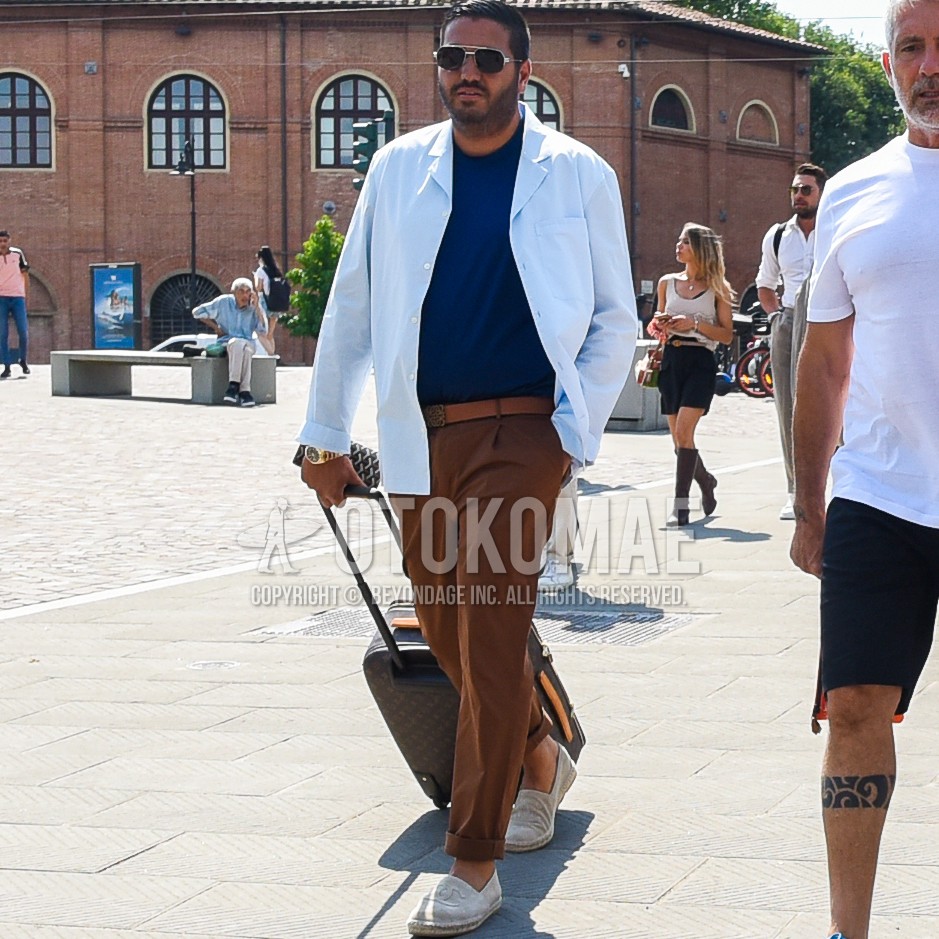 Men's spring summer outfit with silver plain sunglasses, blue plain shirt jacket, navy plain t-shirt, brown plain leather belt, brown plain slacks, white slip-on sneakers, brown small crest suitcase.