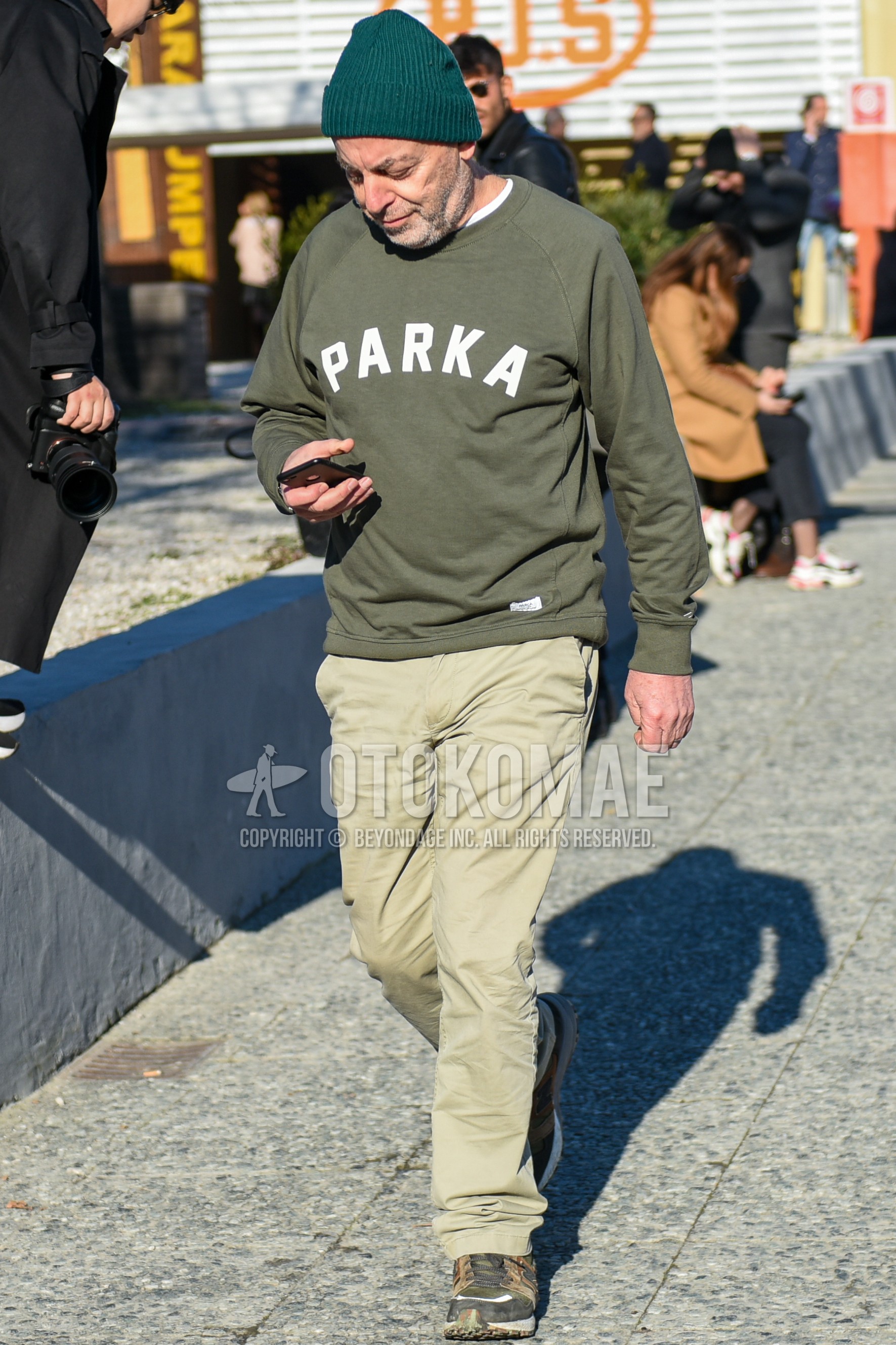 Men's spring autumn outfit with green plain knit cap, olive green deca logo sweatshirt, beige plain chinos, gray low-cut sneakers.