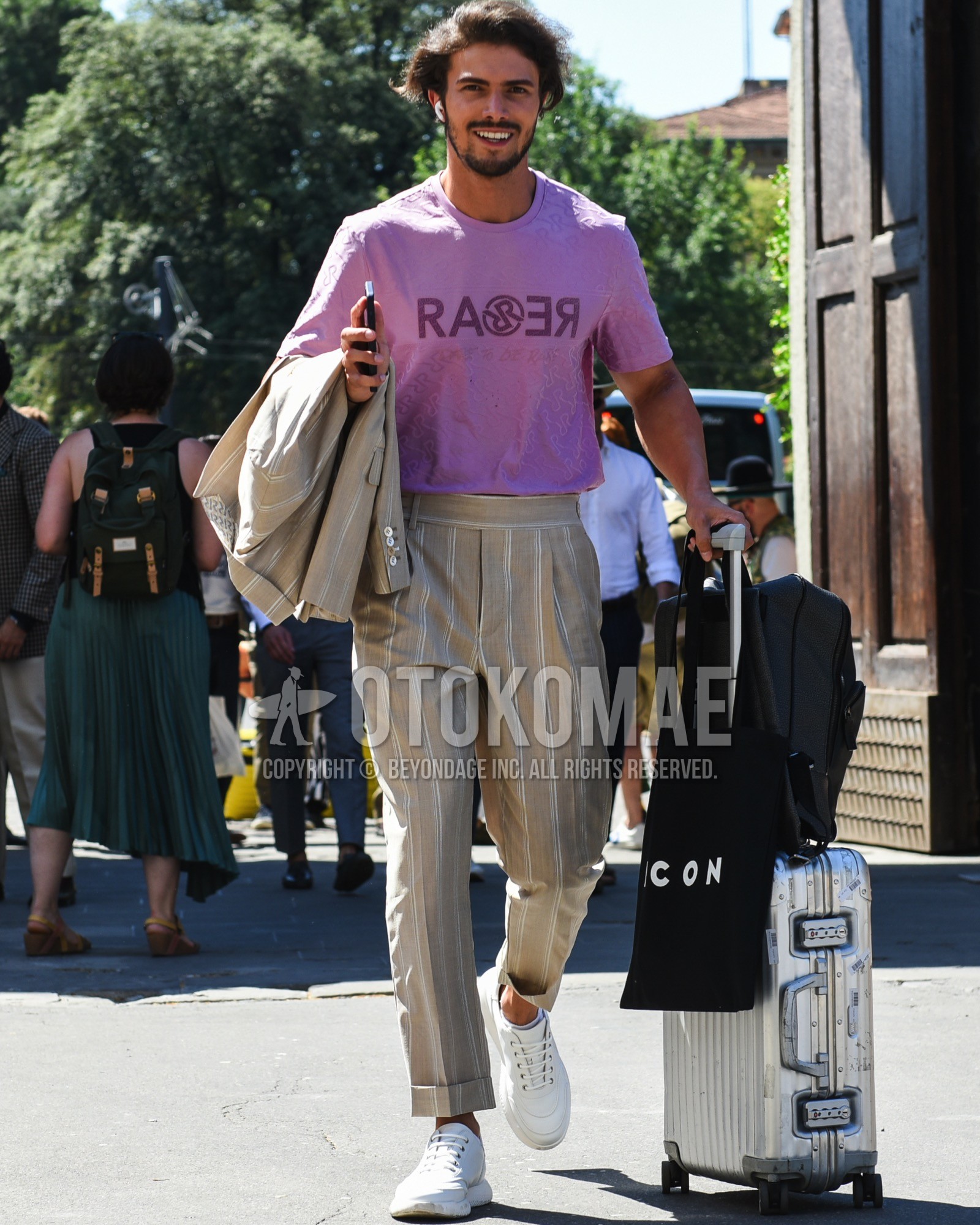 Men's spring summer outfit with pink deca logo t-shirt, white low-cut sneakers, silver plain suitcase, black deca logo tote bag, black plain backpack, beige stripes suit.