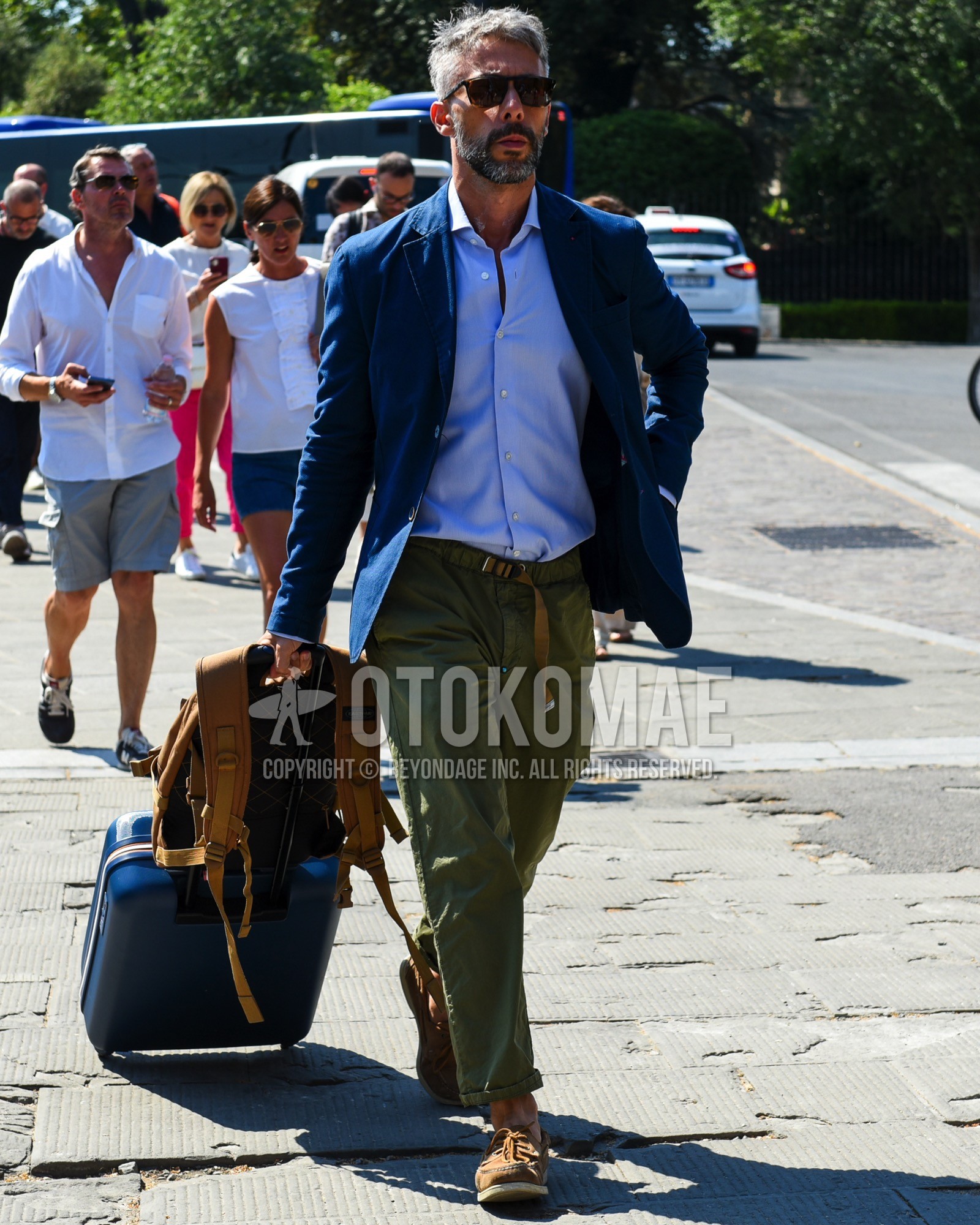 Men's spring summer outfit with brown tortoiseshell sunglasses, navy plain tailored jacket, blue plain shirt, olive green plain bottoms, brown moccasins/deck shoes leather shoes, navy plain suitcase, brown plain backpack.