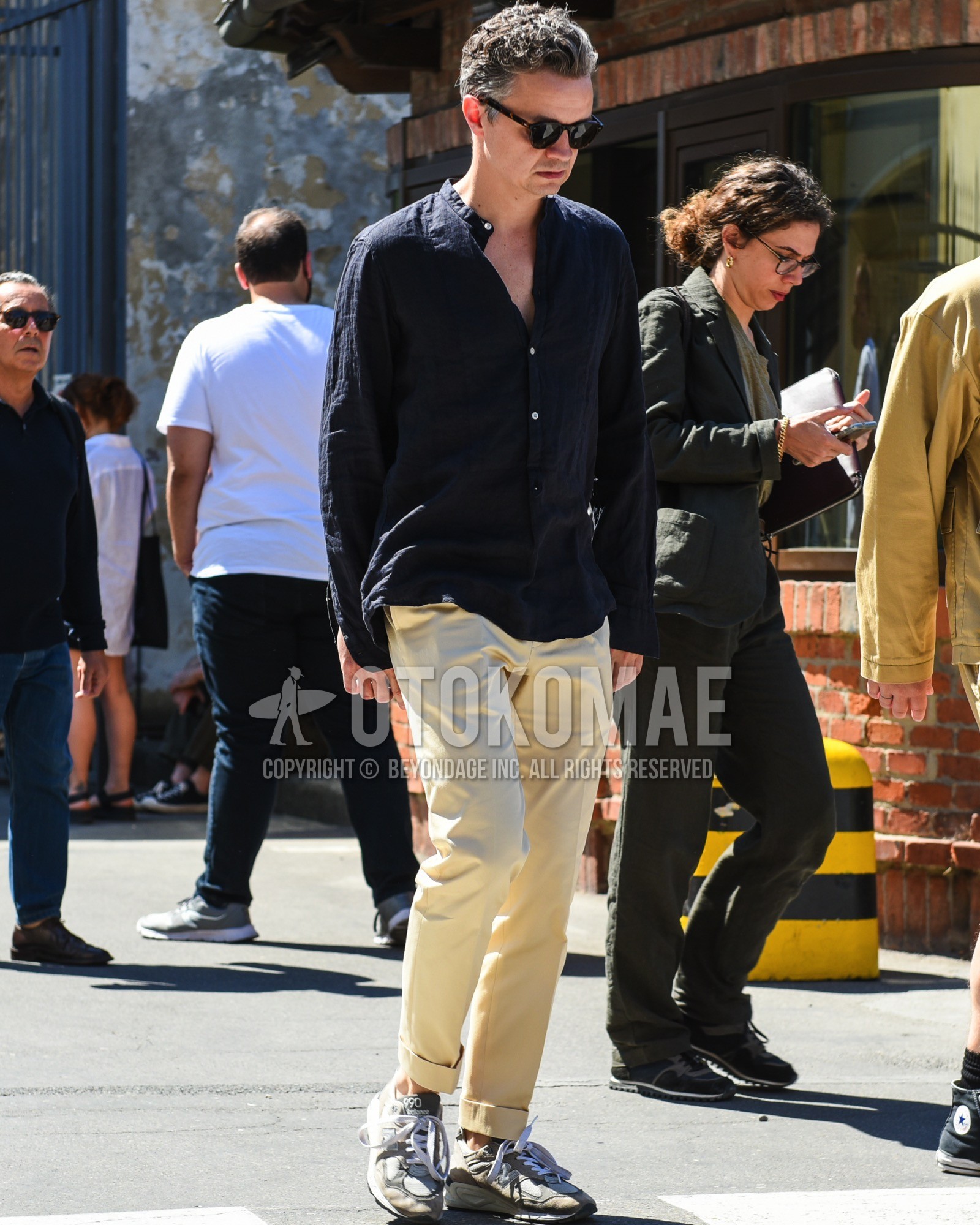 Men's spring summer outfit with black plain sunglasses, black plain shirt, beige plain chinos, gray low-cut sneakers.
