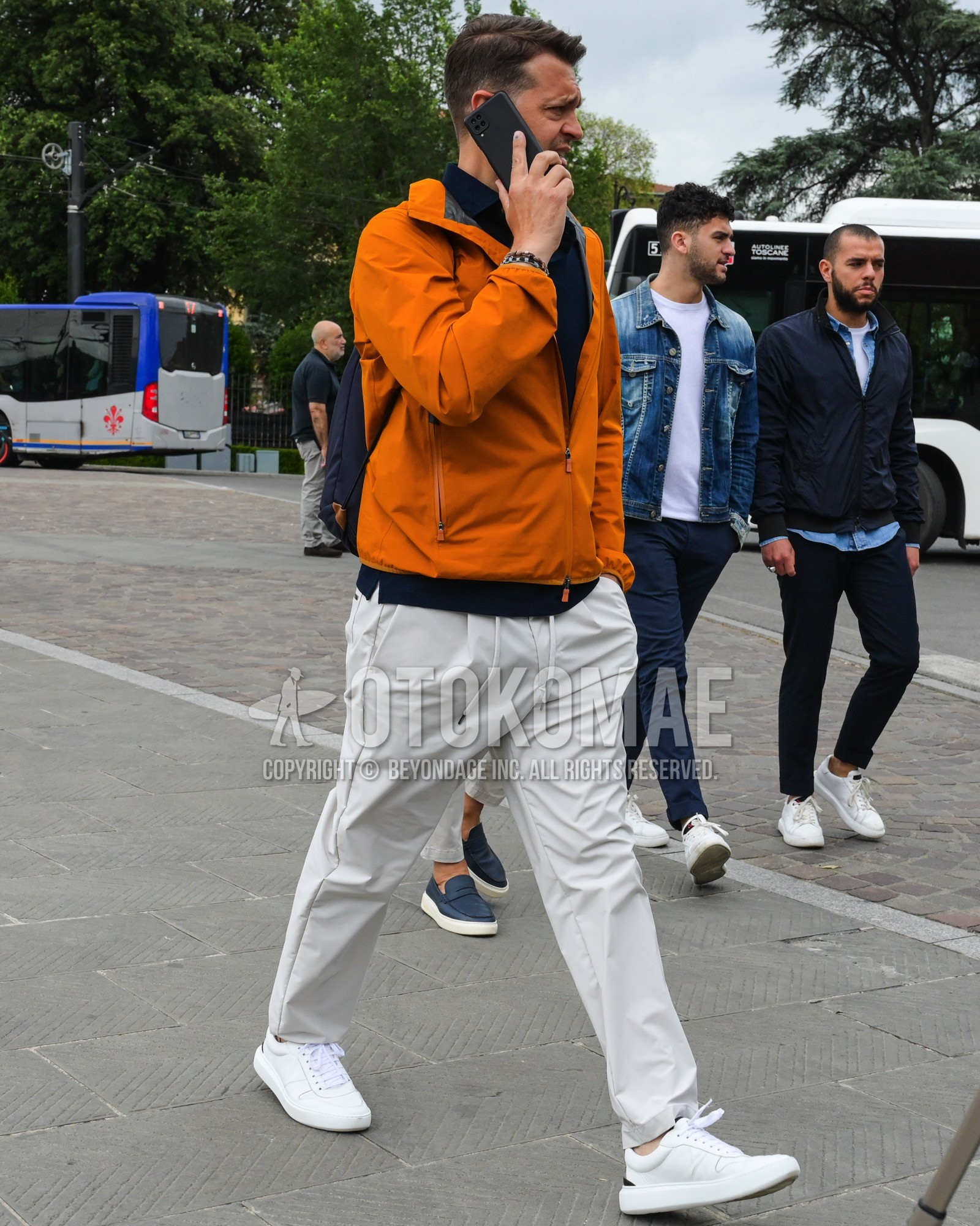 Men's spring summer autumn outfit with orange plain windbreaker, white plain easy pants, white low-cut sneakers, black plain backpack.