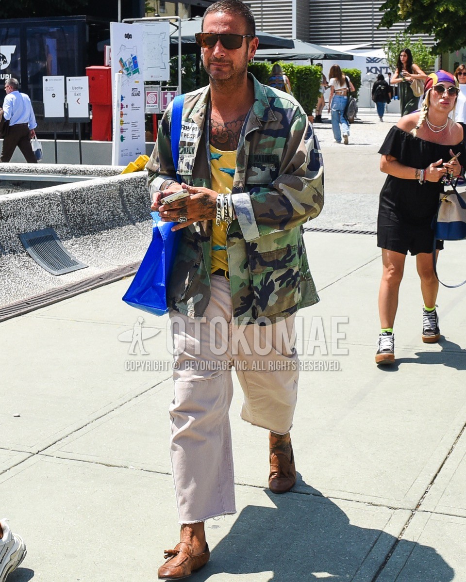 Men's spring summer outfit with brown tortoiseshell sunglasses, olive green camouflage field jacket/hunting jacket, yellow graphic tank top, black plain leather belt, beige plain wide pants, beige plain baker pants, brown tassel loafers leather shoes, blue plain tote bag.