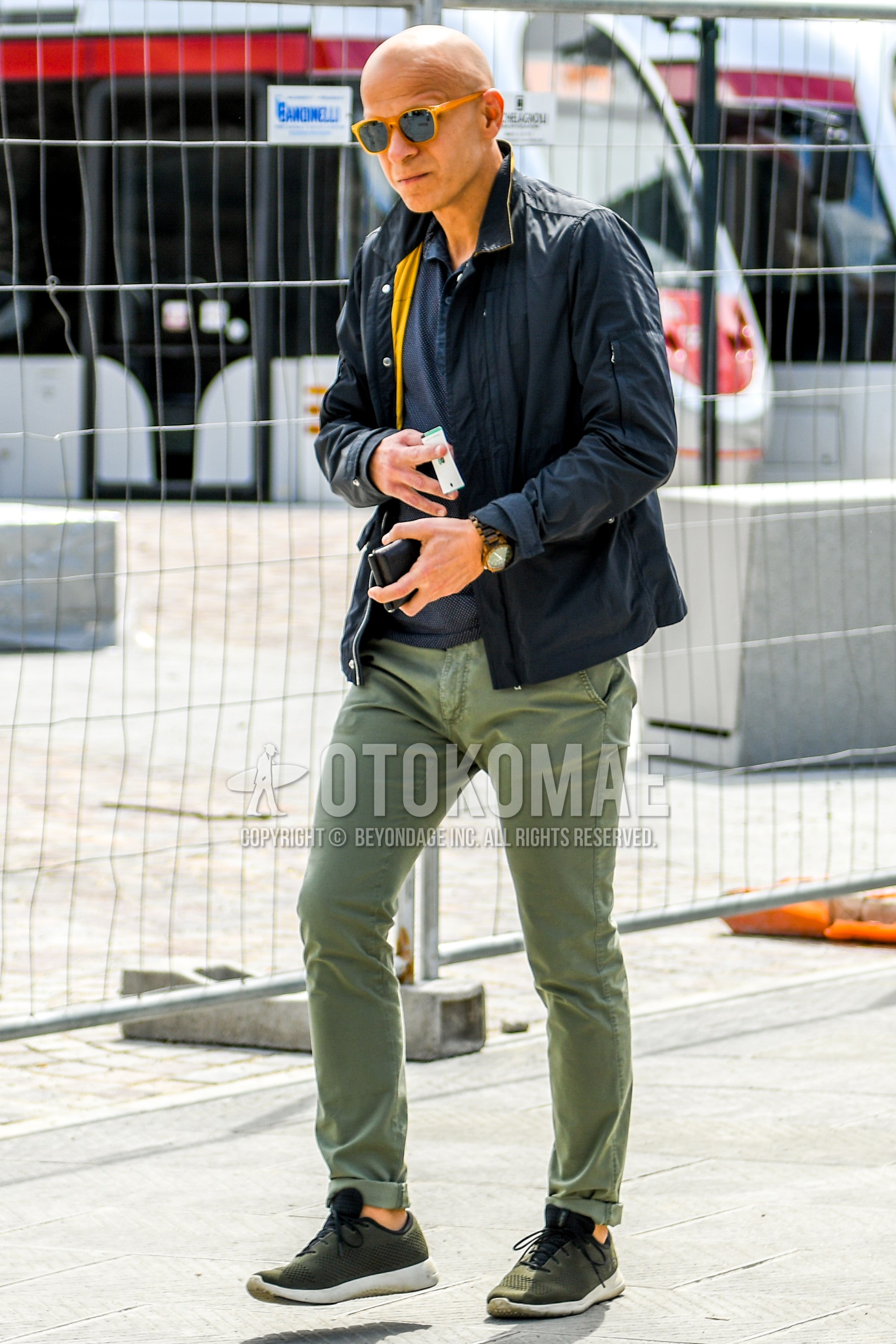 Men's spring autumn outfit with beige plain sunglasses, black plain outerwear, navy dots polo shirt, olive green plain chinos, olive green low-cut sneakers.
