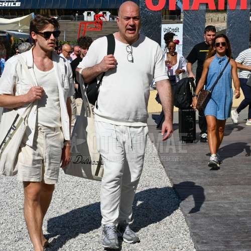 黒無地のメガネ、ニットの白無地のTシャツ、白無地のスラックス、白無地のイージーパンツ、グレーローカットのスニーカー、白レタードのトートバッグ、黒無地のバックパックを合わせた夏春のメンズコーデ・着こなし。