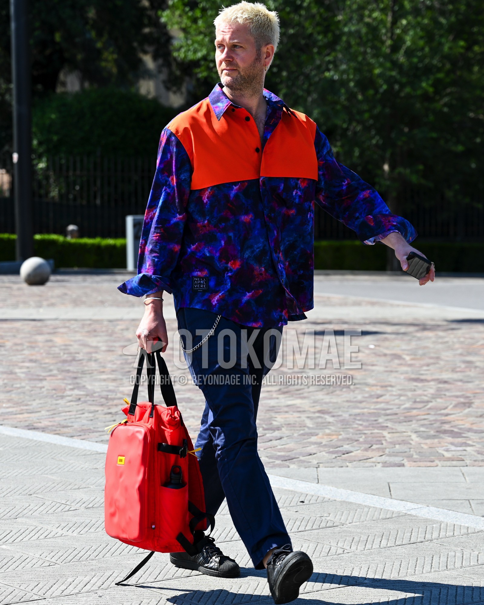 Men's spring summer autumn outfit with purple whole pattern windbreaker, navy plain chinos, black low-cut sneakers, red plain tote bag.