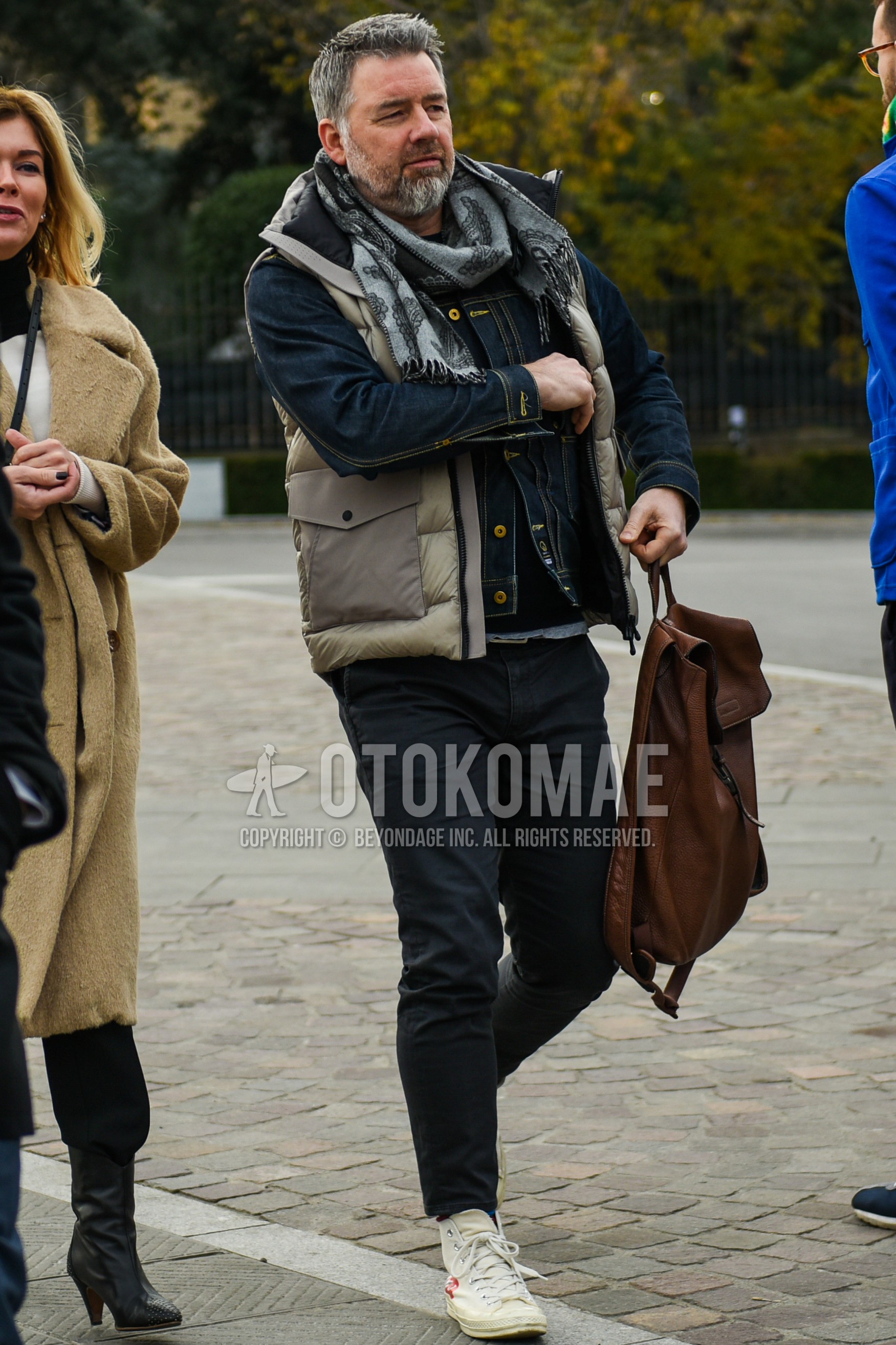Men's autumn winter outfit with gray scarf scarf, beige outerwear down jacket, navy plain denim jacket, beige plain casual vest, black plain chinos, white high-cut sneakers, brown plain backpack.