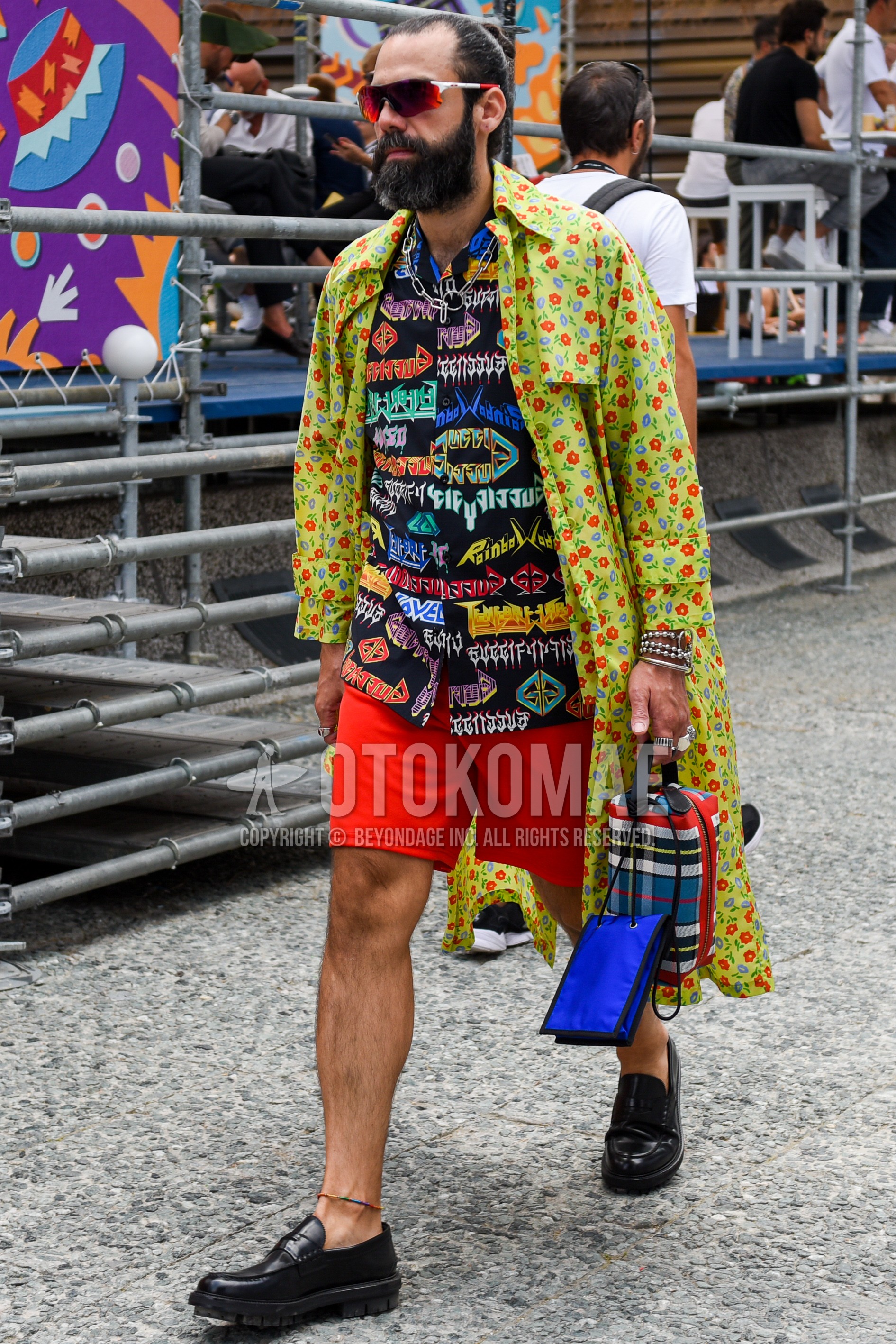 Men's spring summer autumn outfit with white red plain sunglasses, yellow plain trench coat, multi-color graphic shirt, red plain short pants, black coin loafers leather shoes.