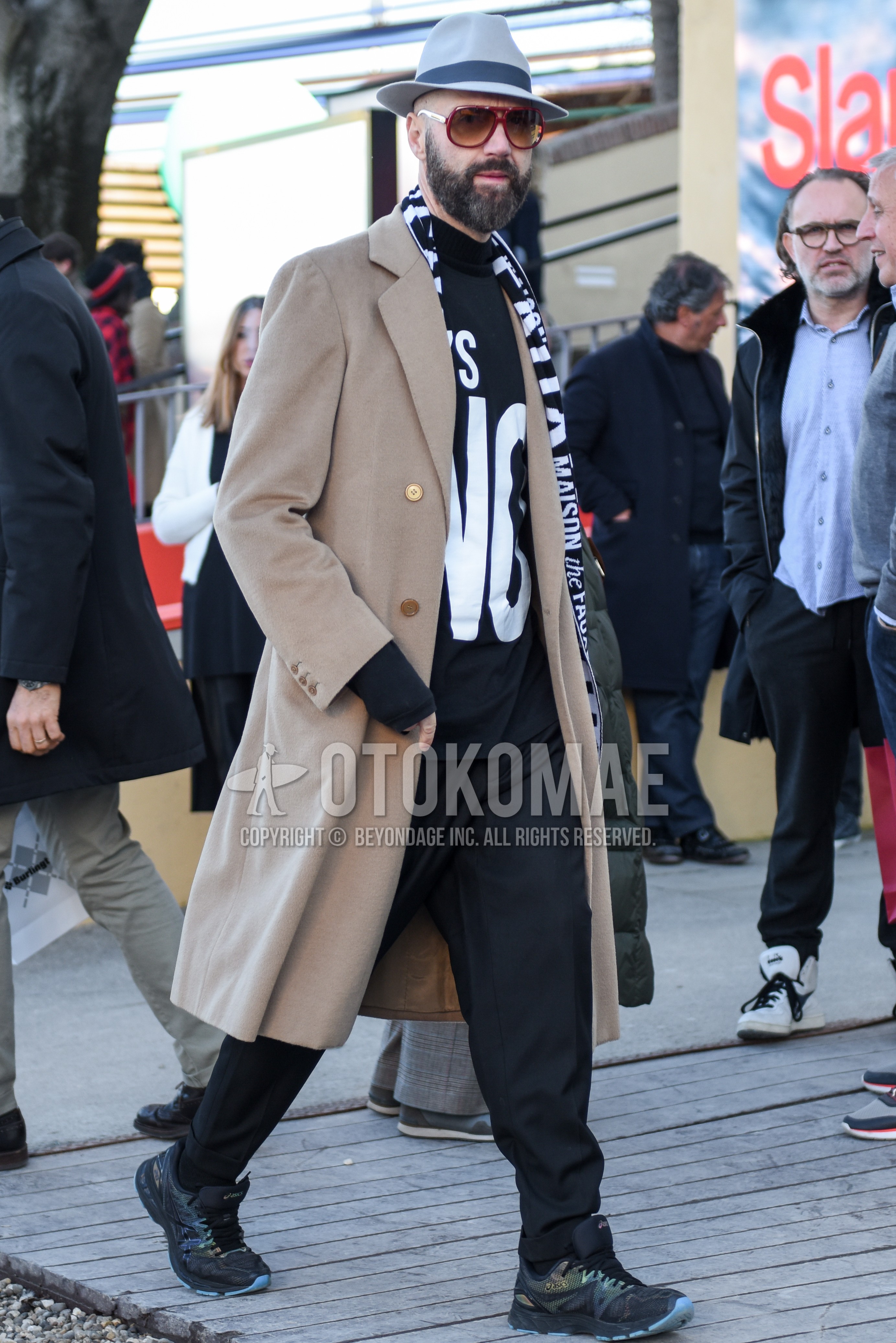 Men's autumn winter outfit with gray plain hat, brown tortoiseshell sunglasses, black scarf scarf, beige plain chester coat, black graphic t-shirt, gray plain slacks, black low-cut sneakers.
