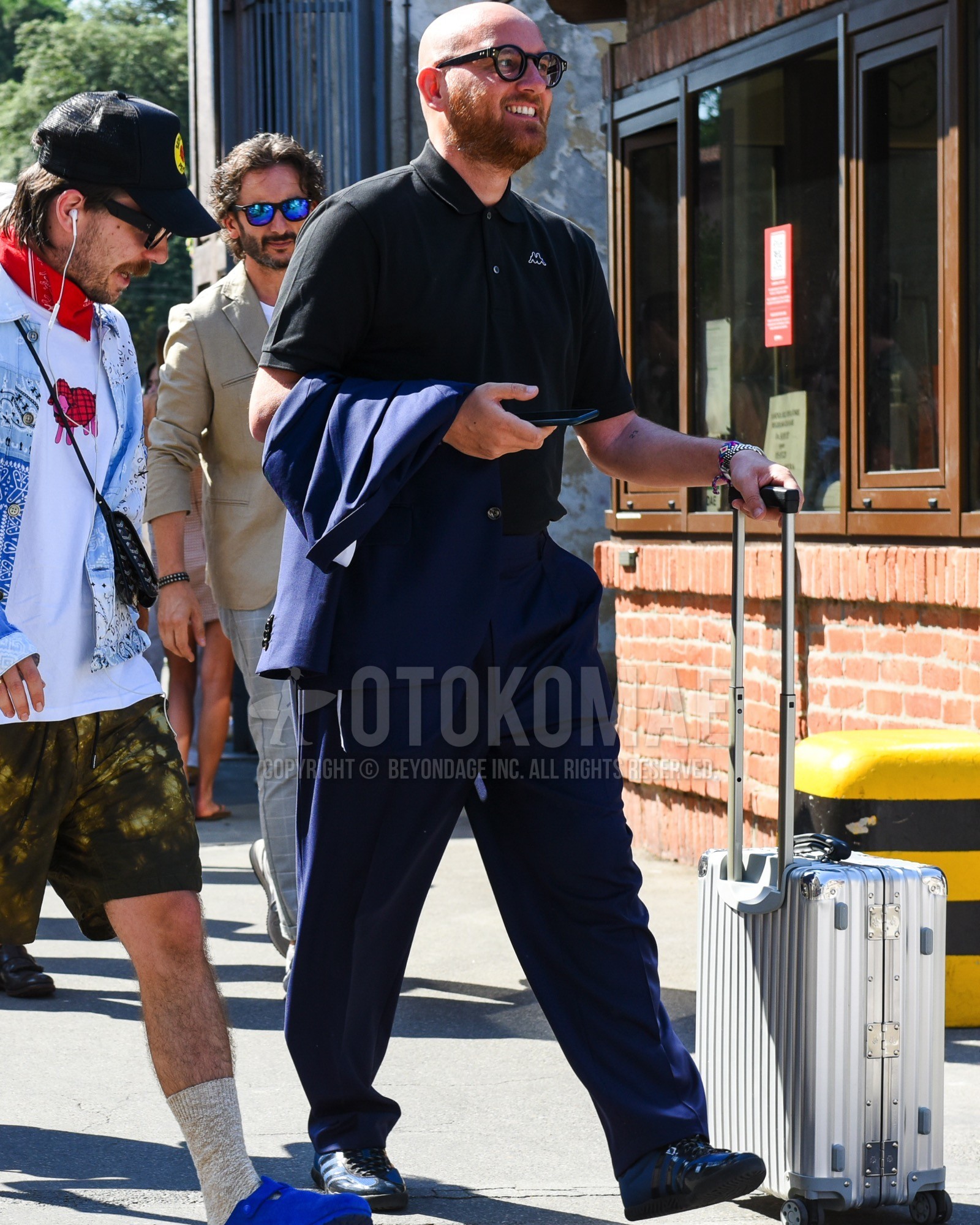 Men's spring summer outfit with black plain glasses, black one point polo shirt, navy low-cut sneakers, silver plain suitcase, navy plain suit.