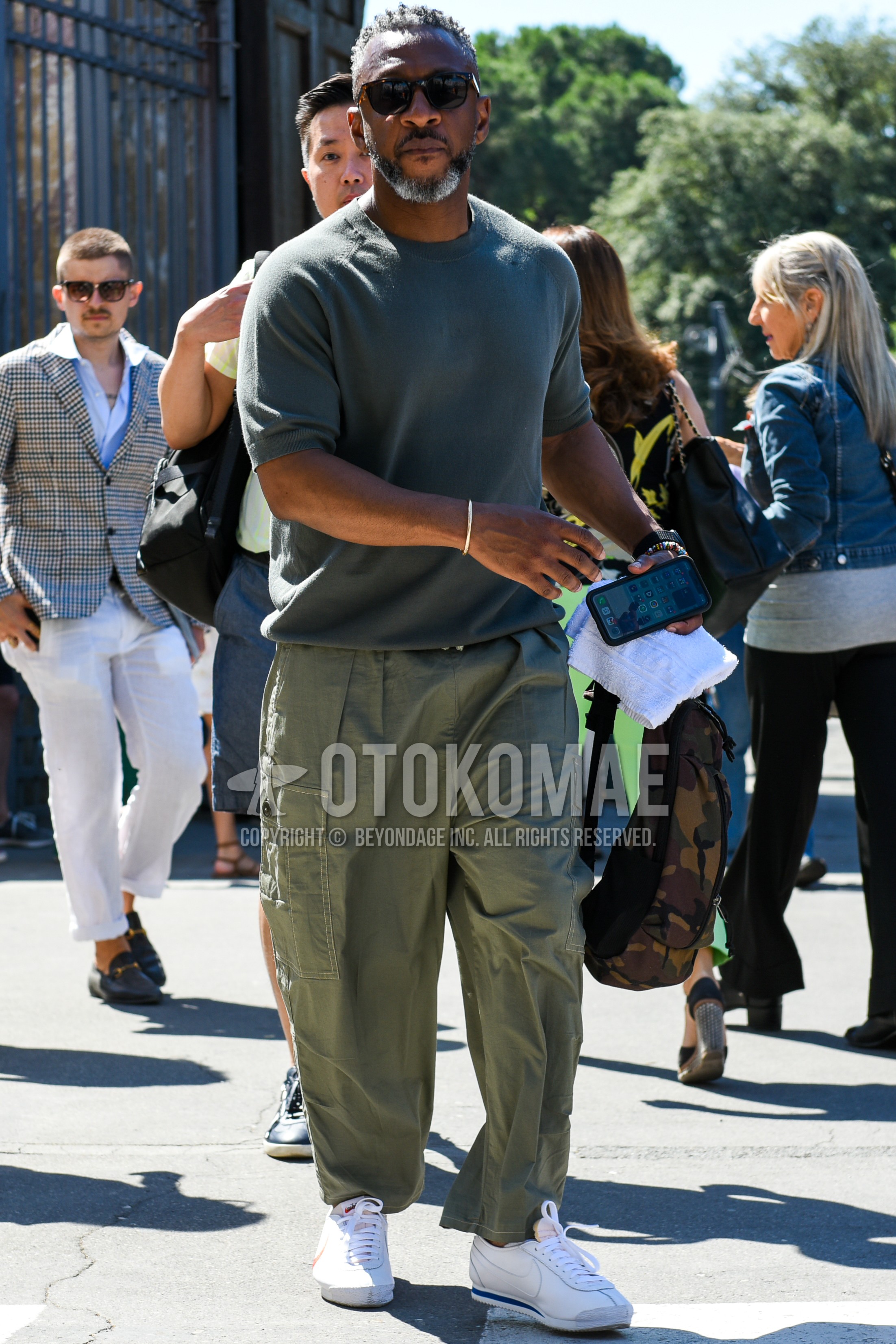 Men's spring summer outfit with black plain sunglasses, olive green plain t-shirt, olive green plain cargo pants, white low-cut sneakers, olive green camouflage backpack.
