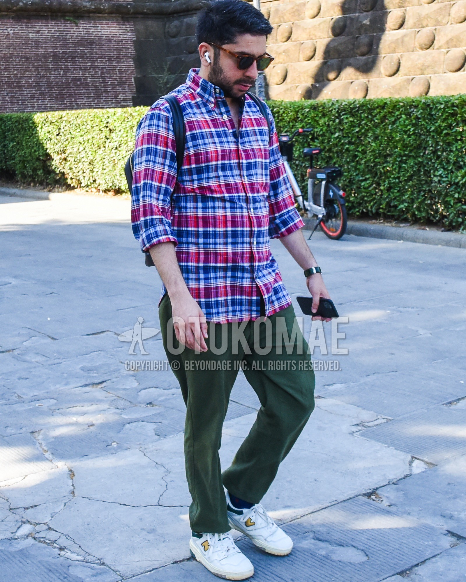 Men's spring summer outfit with brown tortoiseshell sunglasses, blue red check shirt, olive green plain cotton pants, white low-cut sneakers, black plain backpack.